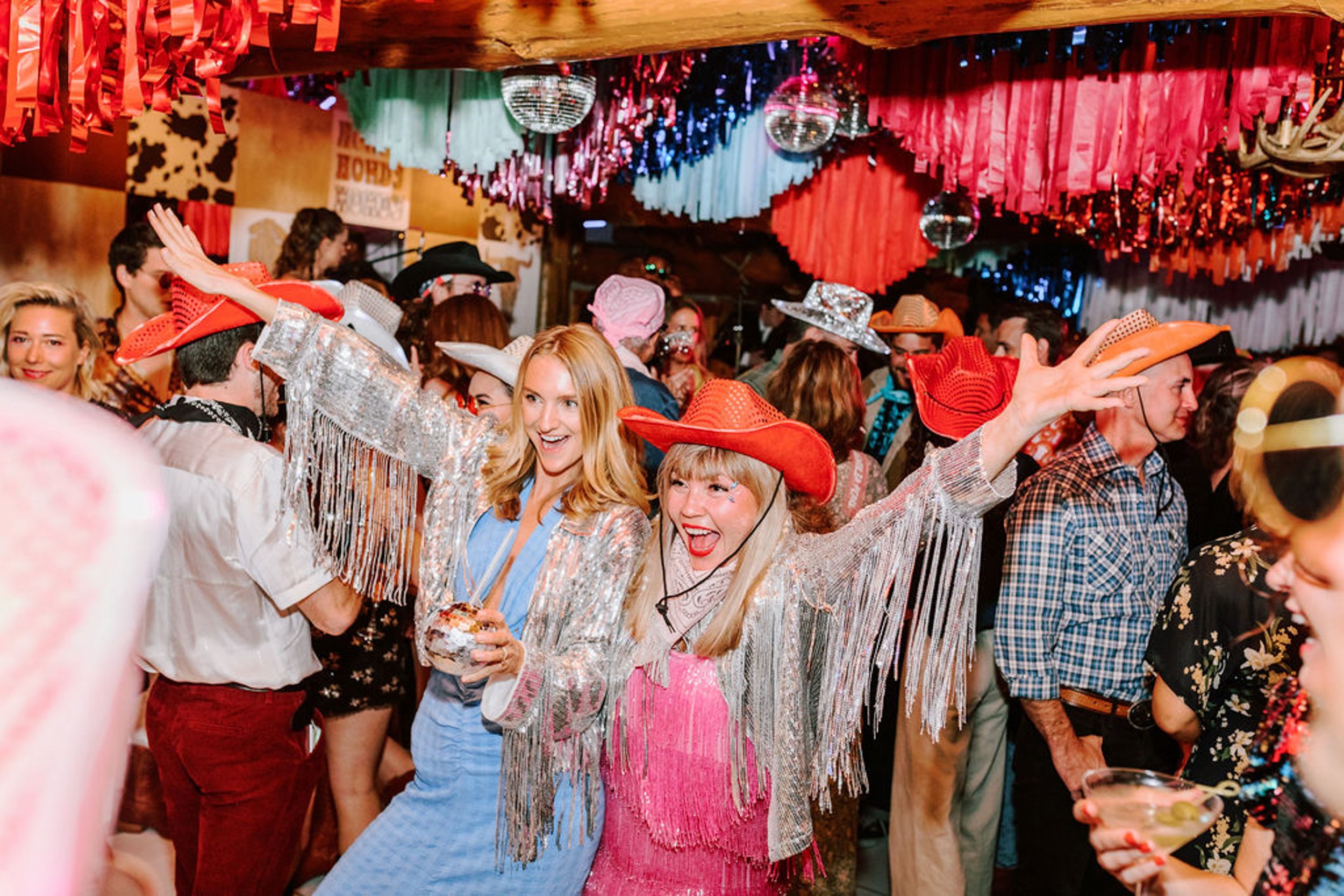 Women in silver fringe jackets and red cowboy hats dancing and celebrating at Pine Creek Cookhouse wedding reception with disco balls and colorful streamers overhead