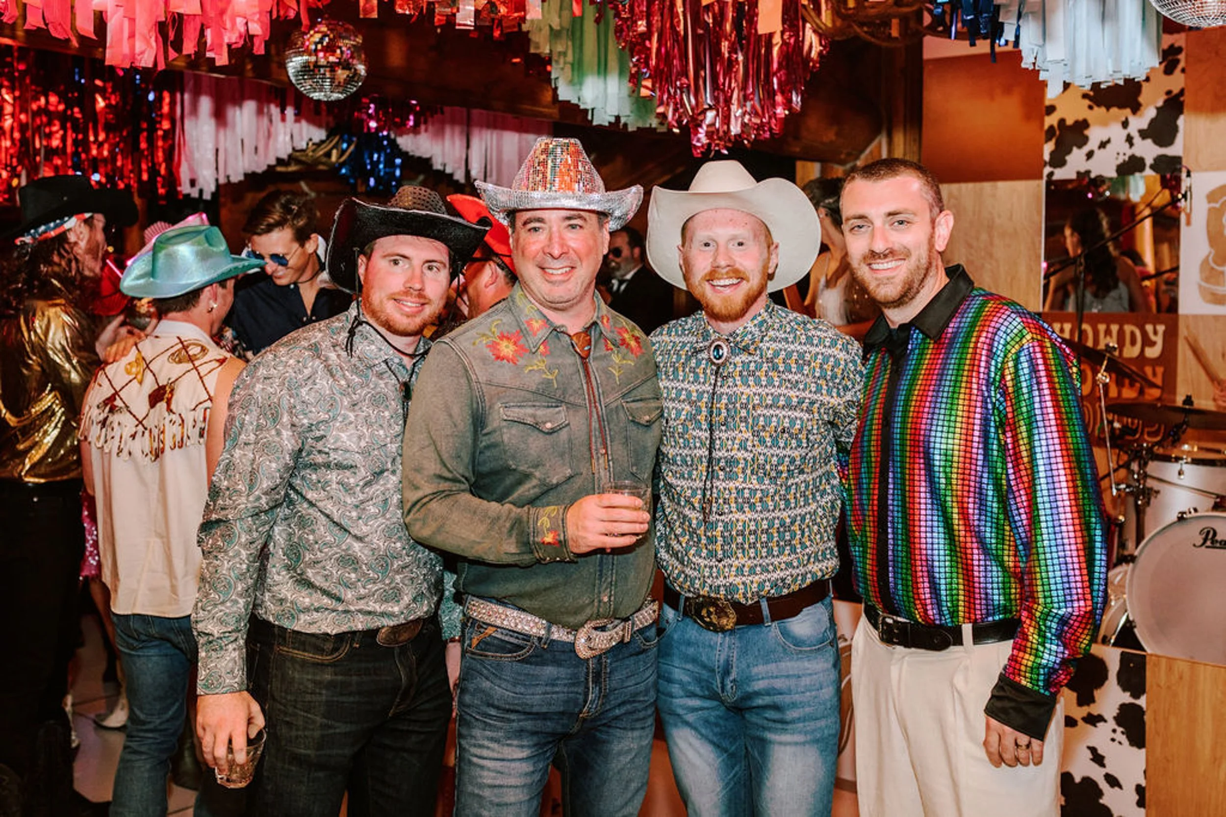 Four male wedding guests in patterned western shirts and cowboy hats including one in a rainbow sequin shirt at Pine Creek Cookhouse reception