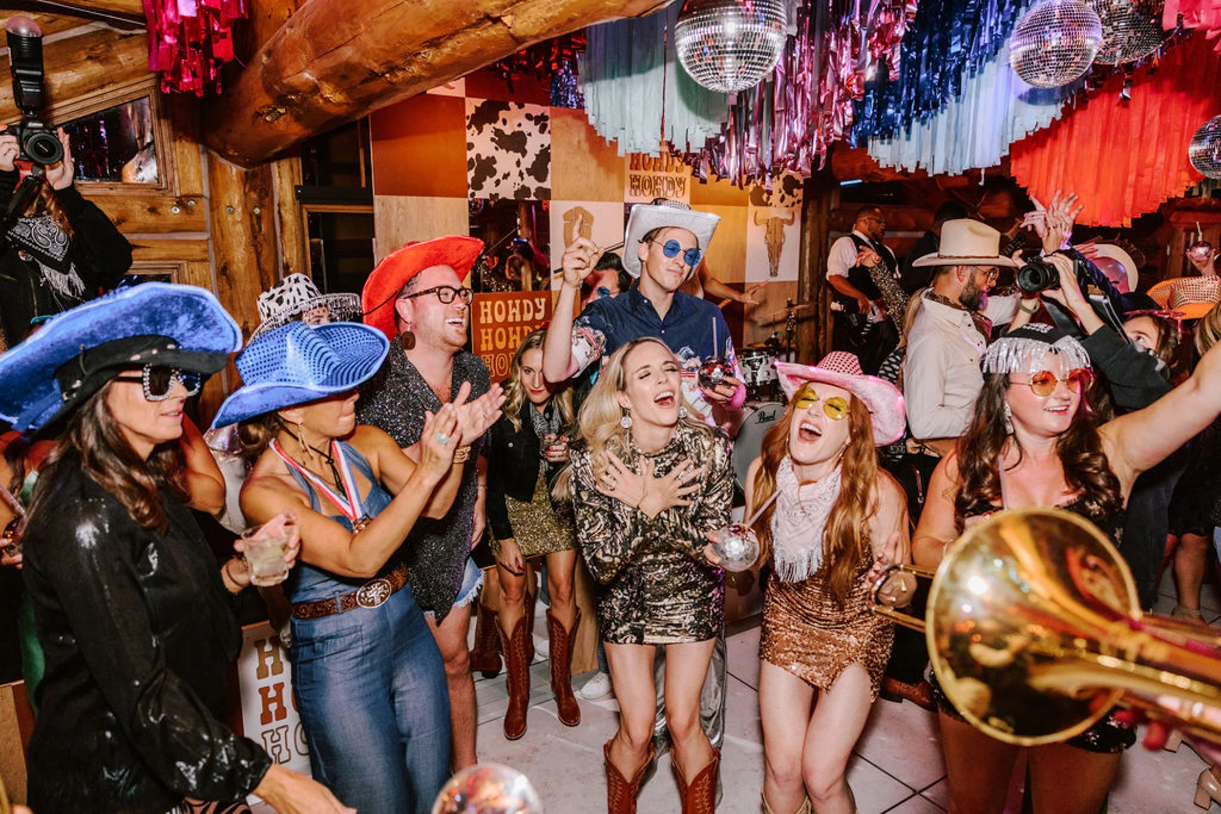 Wedding guests in western and sequin outfits dancing and celebrating at Pine Creek Cookhouse reception with disco balls overhead and live brass instrument player