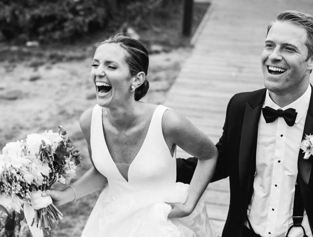 A black-and-white photo of a smiling bride and groom walking outdoors, sharing a joyful moment. The bride is holding a bouquet of flowers, and the groom is dressed in a tuxedo with a bow tie.