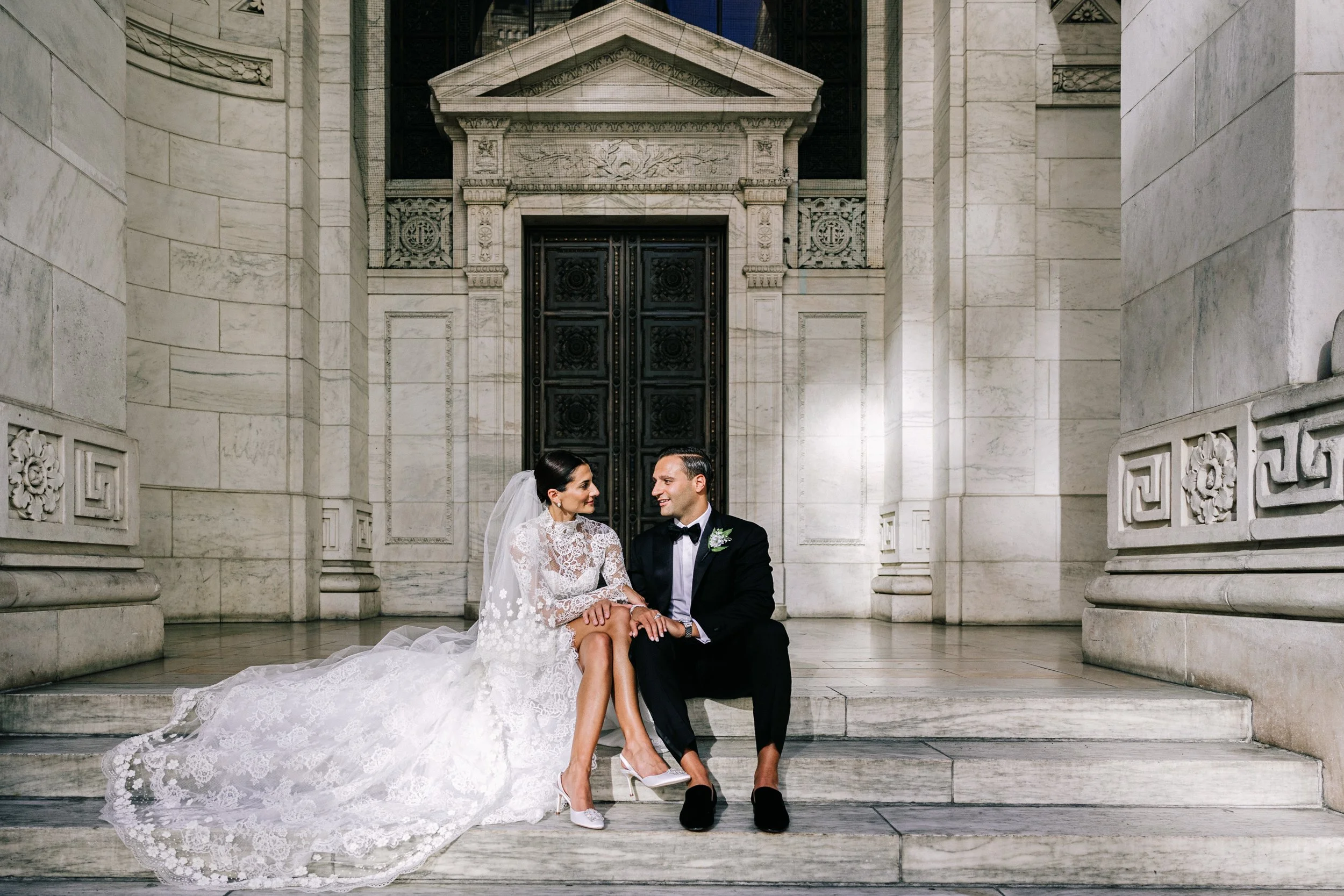 Bride and groom sitting on the exterior steps of the New York Public Library with the ornate columns and bronze doors behind them