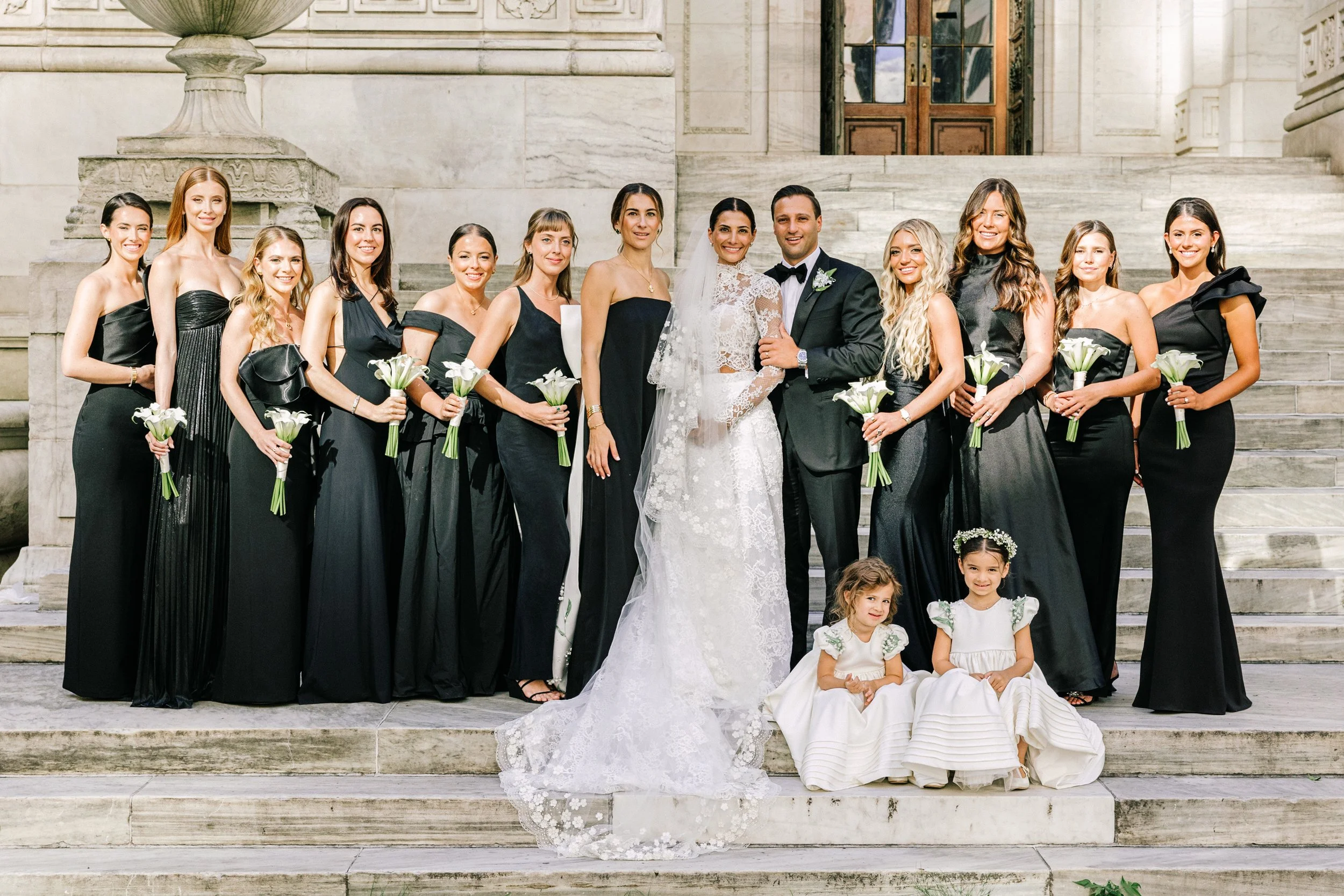 Bride with her bridesmaids in black gowns and flower girls in white on the steps of the New York Public Library, NYC wedding