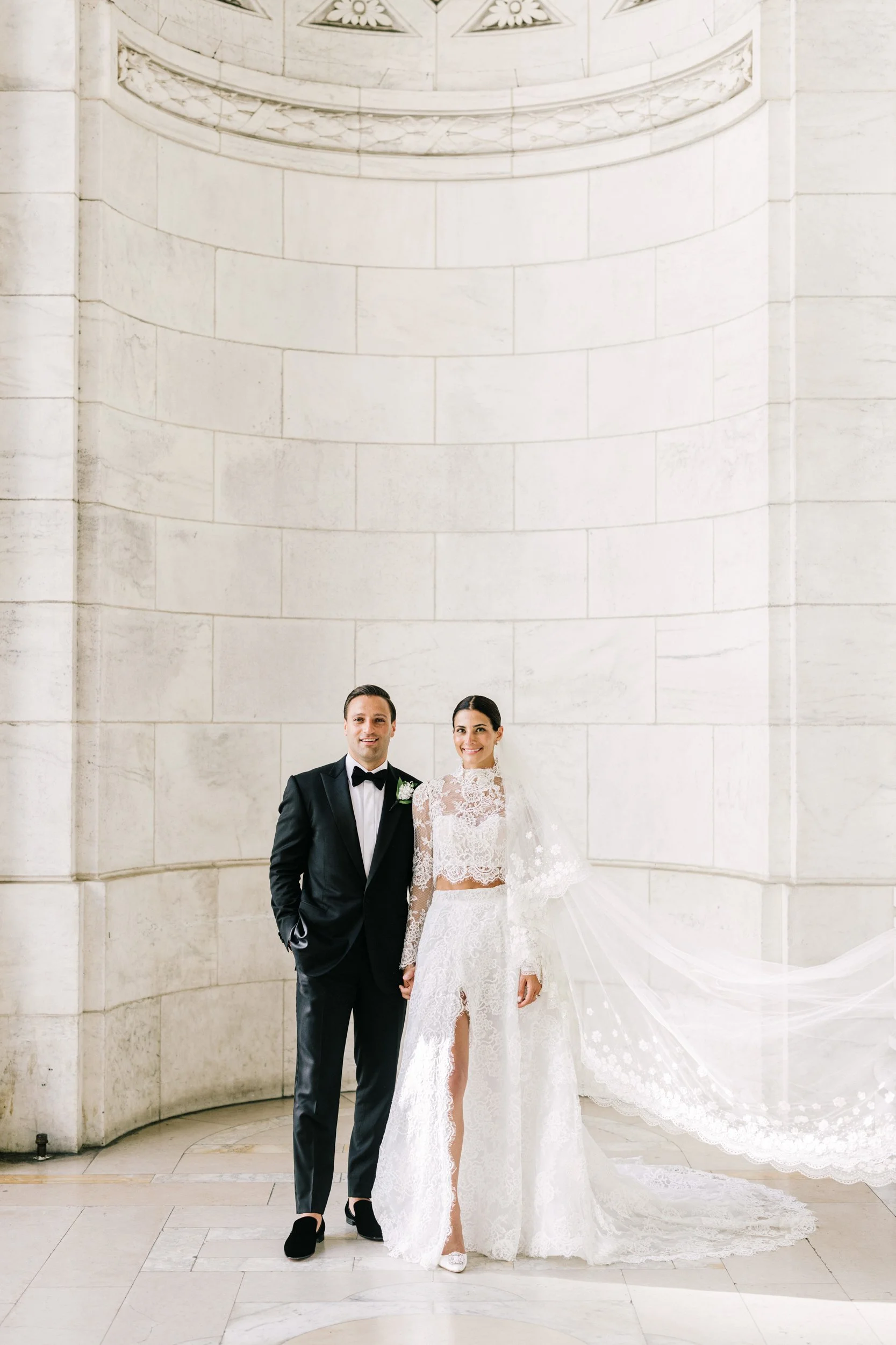 Bride and groom standing together in a full-length portrait against the marble columns of the New York Public Library, New York City wedding