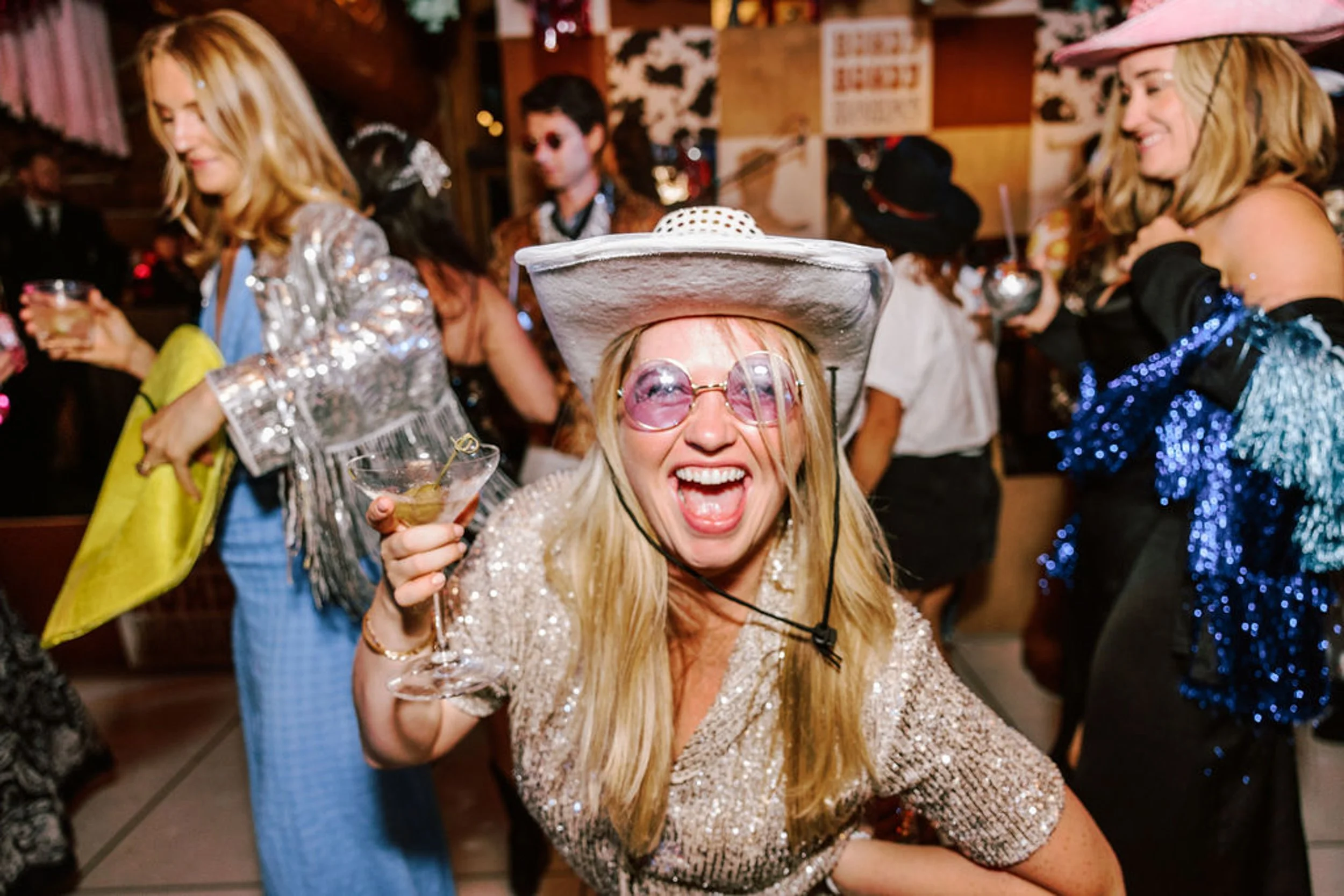 Exuberant female wedding guest in white cowboy hat pink sunglasses and silver sequin top raising a champagne glass at Pine Creek Cookhouse rodeo disco reception
