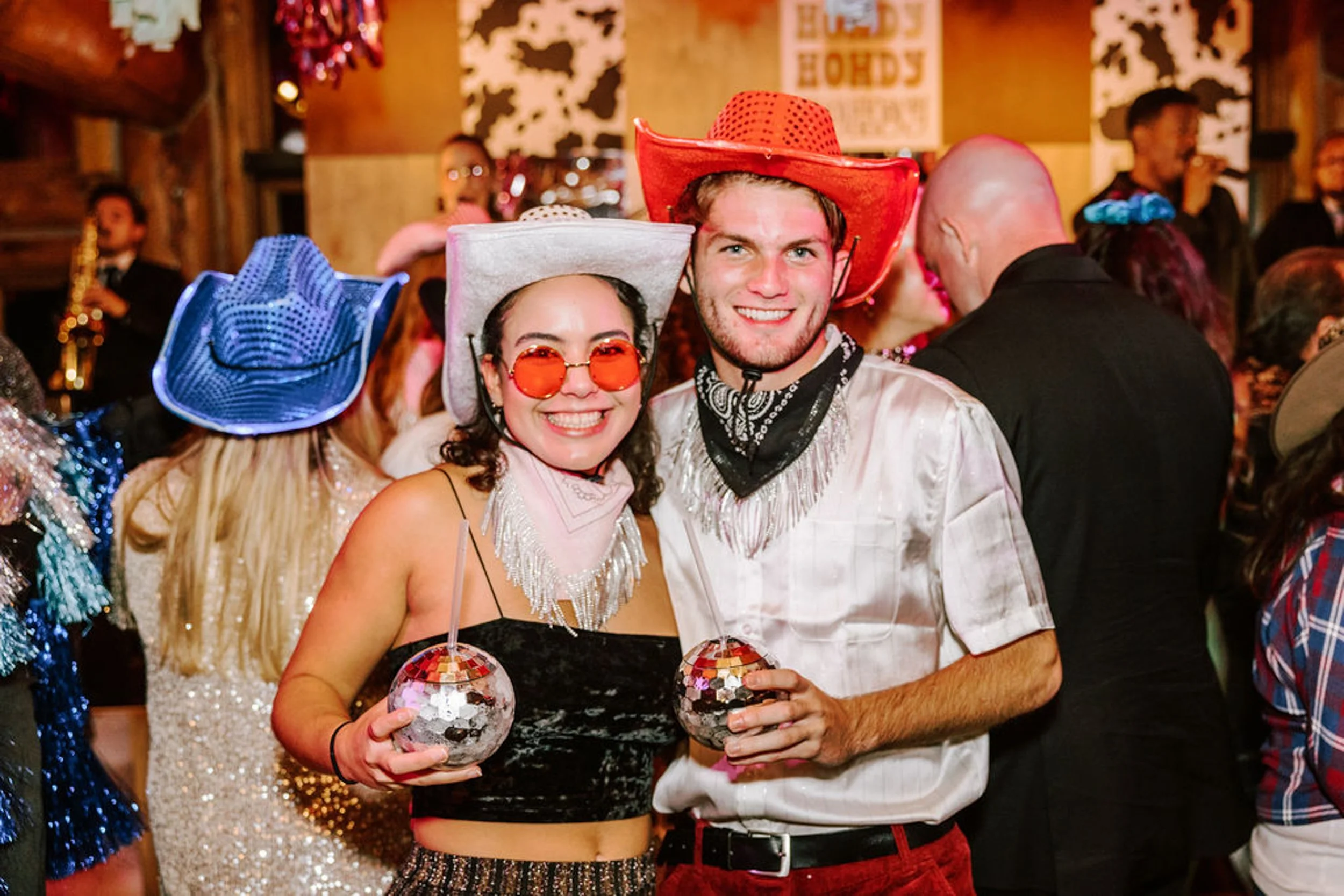 Two wedding guests holding mini disco balls in front of the Howdy Howdy backdrop at Pine Creek Cookhouse reception one in orange glasses and white cowboy hat one in red cowboy hat