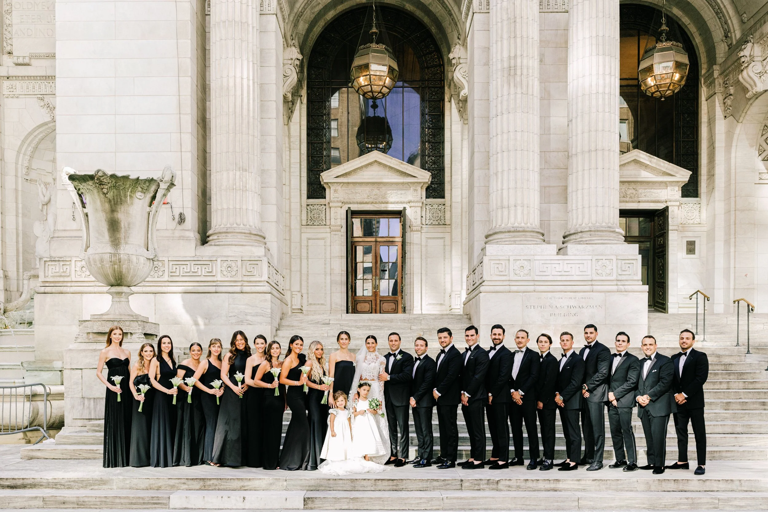 Full wedding party group portrait on the grand steps of the New York Public Library with bridesmaids in black and groomsmen in tuxedos, NYC wedding