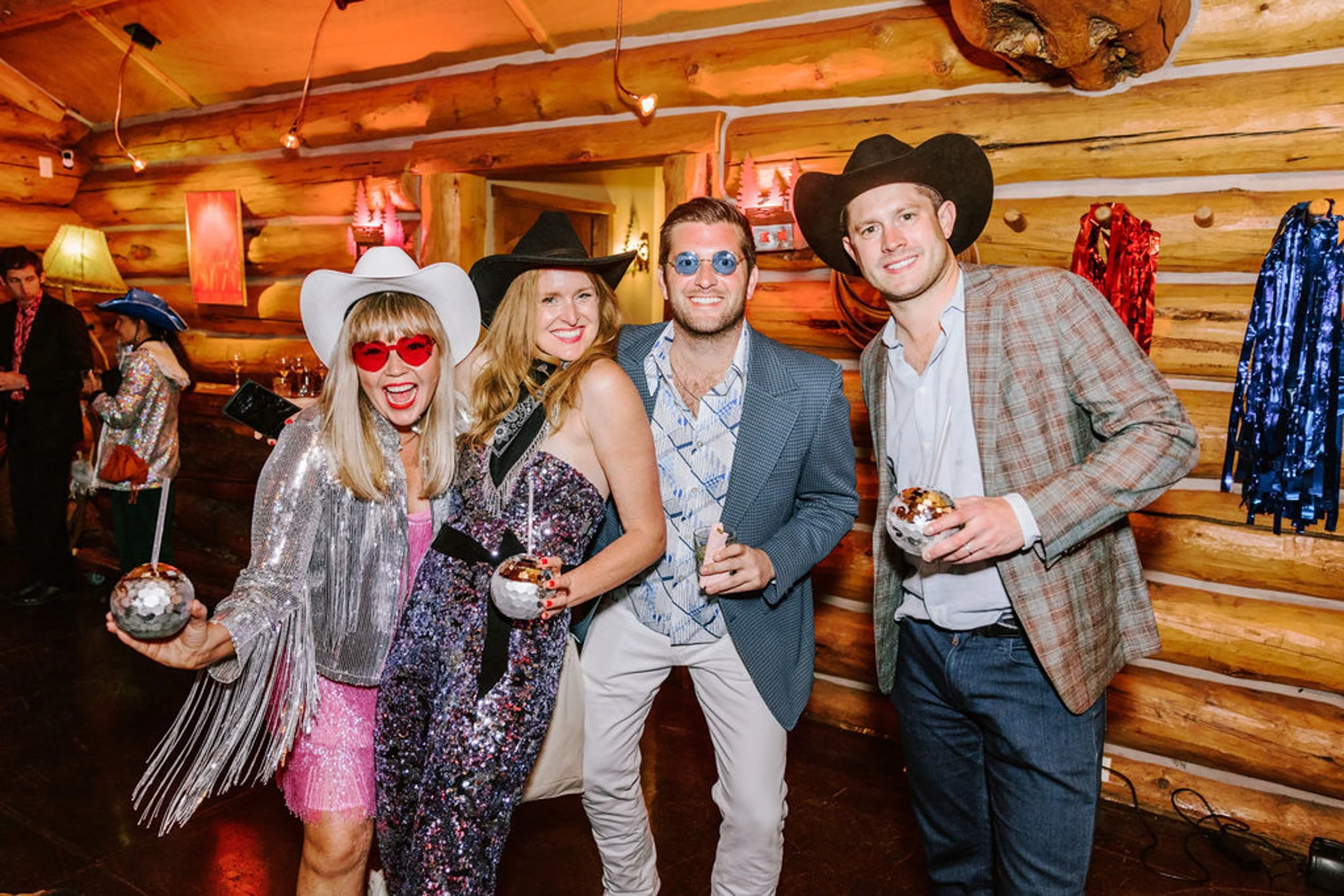 Four wedding guests in western and sequin outfits with cowboy hats holding mini disco balls inside Pine Creek Cookhouse log cabin reception
