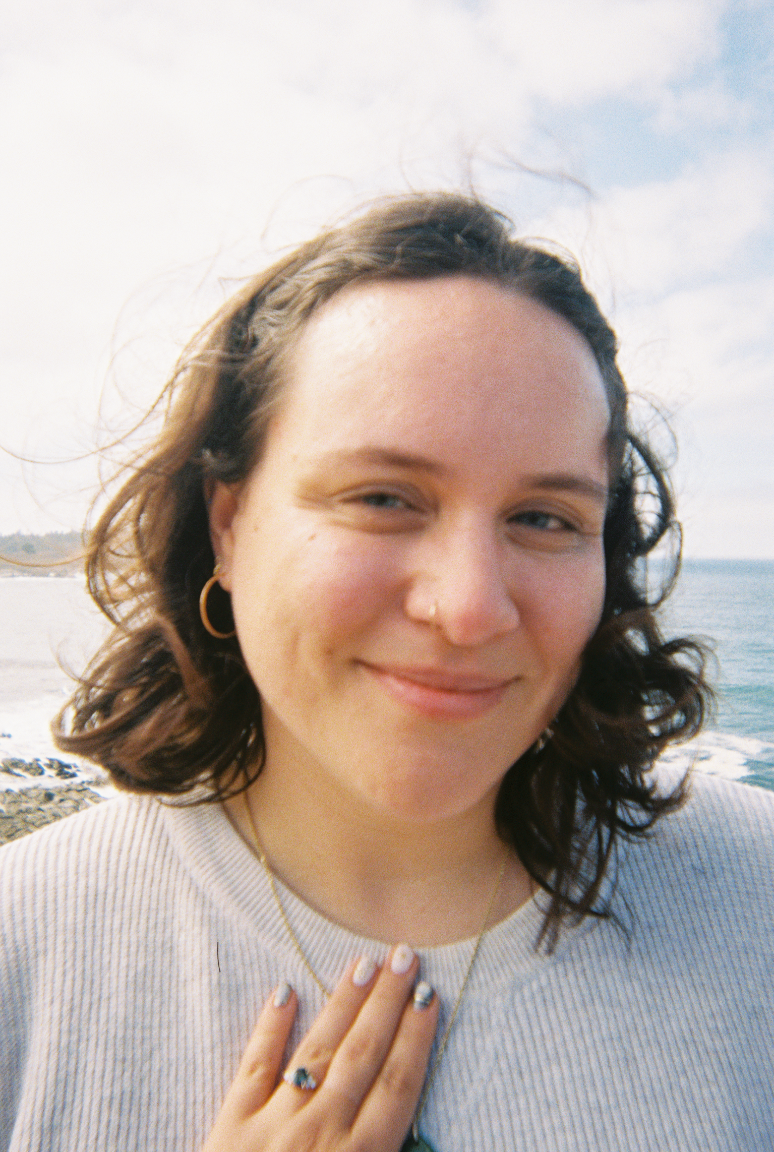 A woman with shoulder-length wavy brown hair, wearing gold hoop earrings and a necklace, smiling at the camera, standing near the ocean with a cloudy sky in the background.