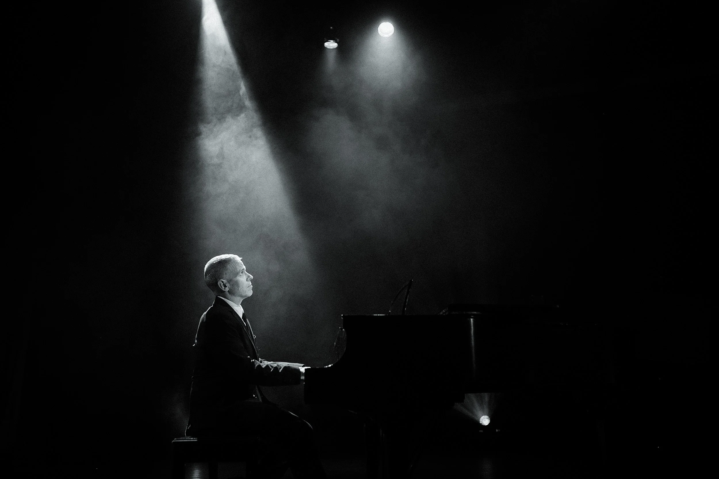A black and white photo of a man playing a grand piano on stage, illuminated by a spotlight from above.