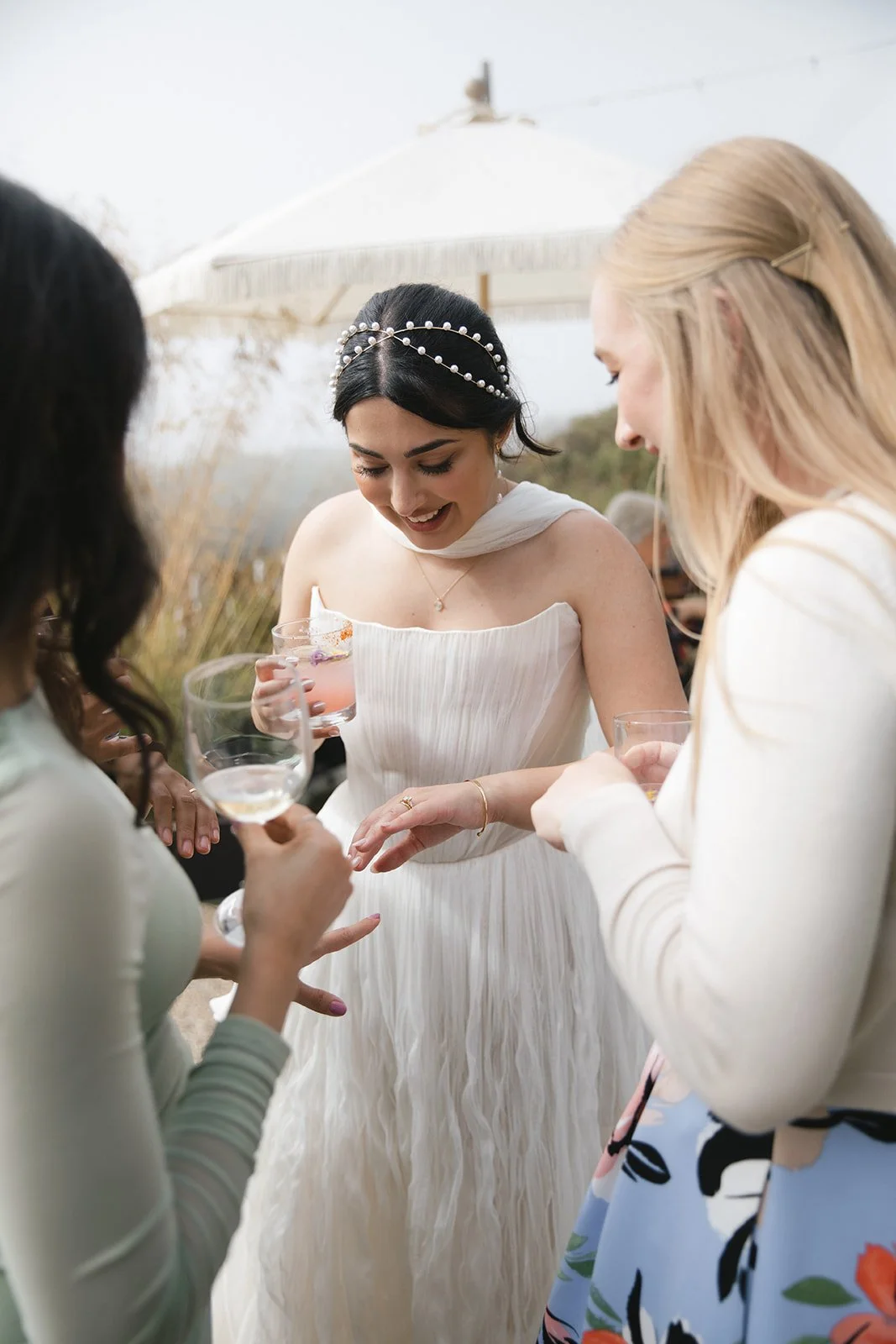 A bride in a white strapless dress with a pearl headband smiling and talking to her guests at an outdoor wedding reception, holding a drink. Guests are gathered around her with drinks in hand, in front of a large outdoor umbrella.