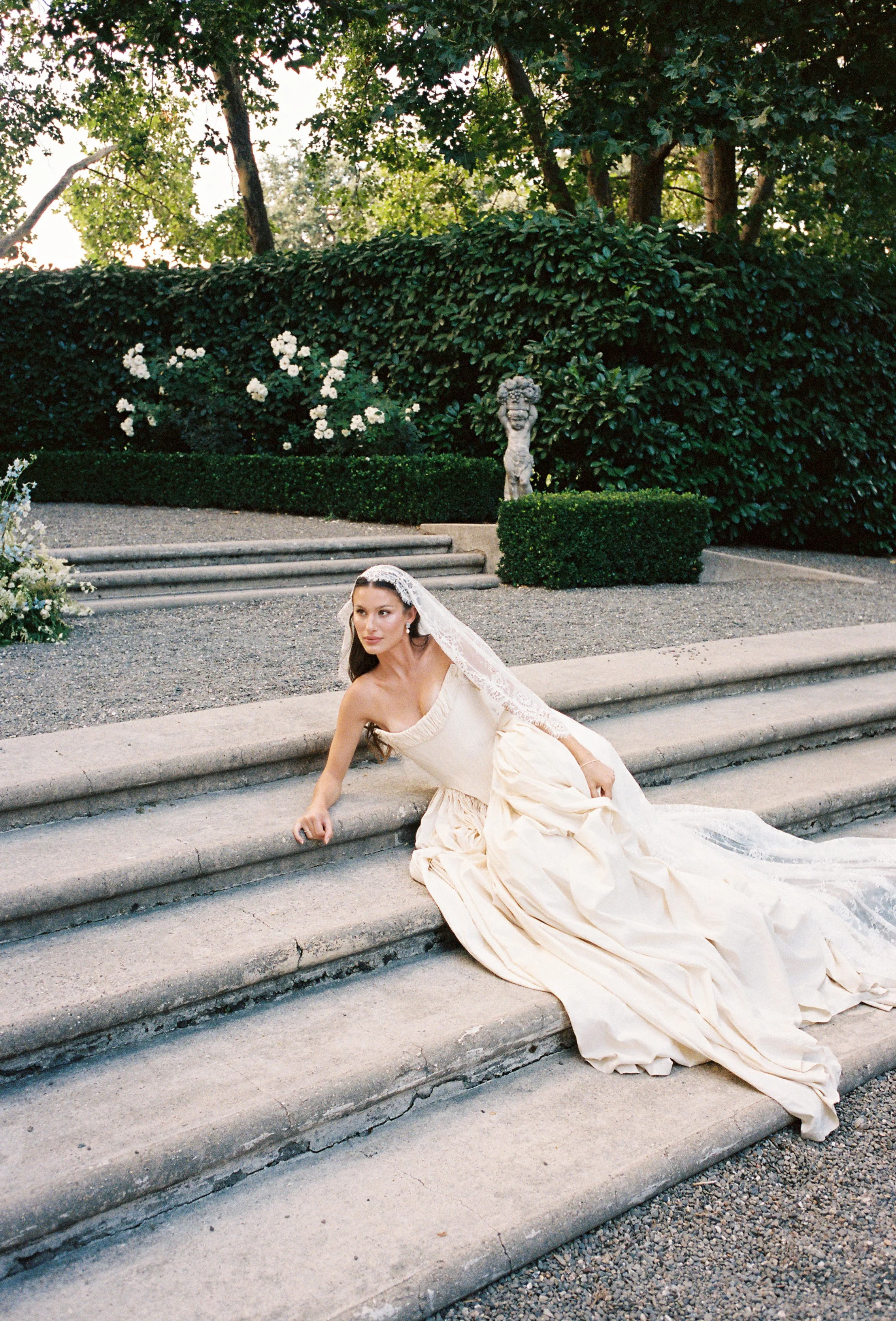 Bride in a wedding dress and veil sitting on outdoor stone stairs with garden shrubbery, trees, and a decorative statue in the background.