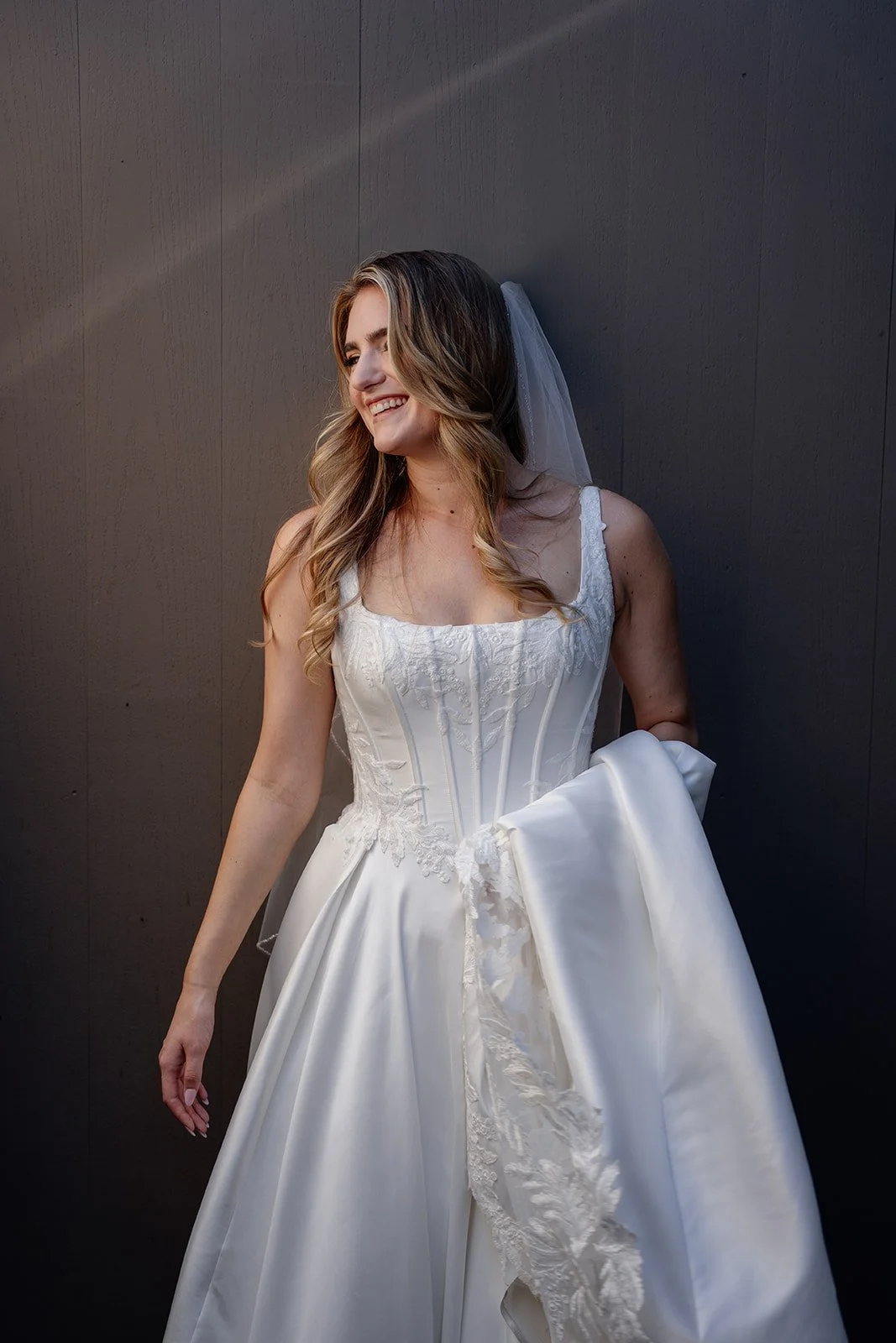 A smiling woman in a wedding dress holding the skirt of her gown, standing against a dark gray wall.