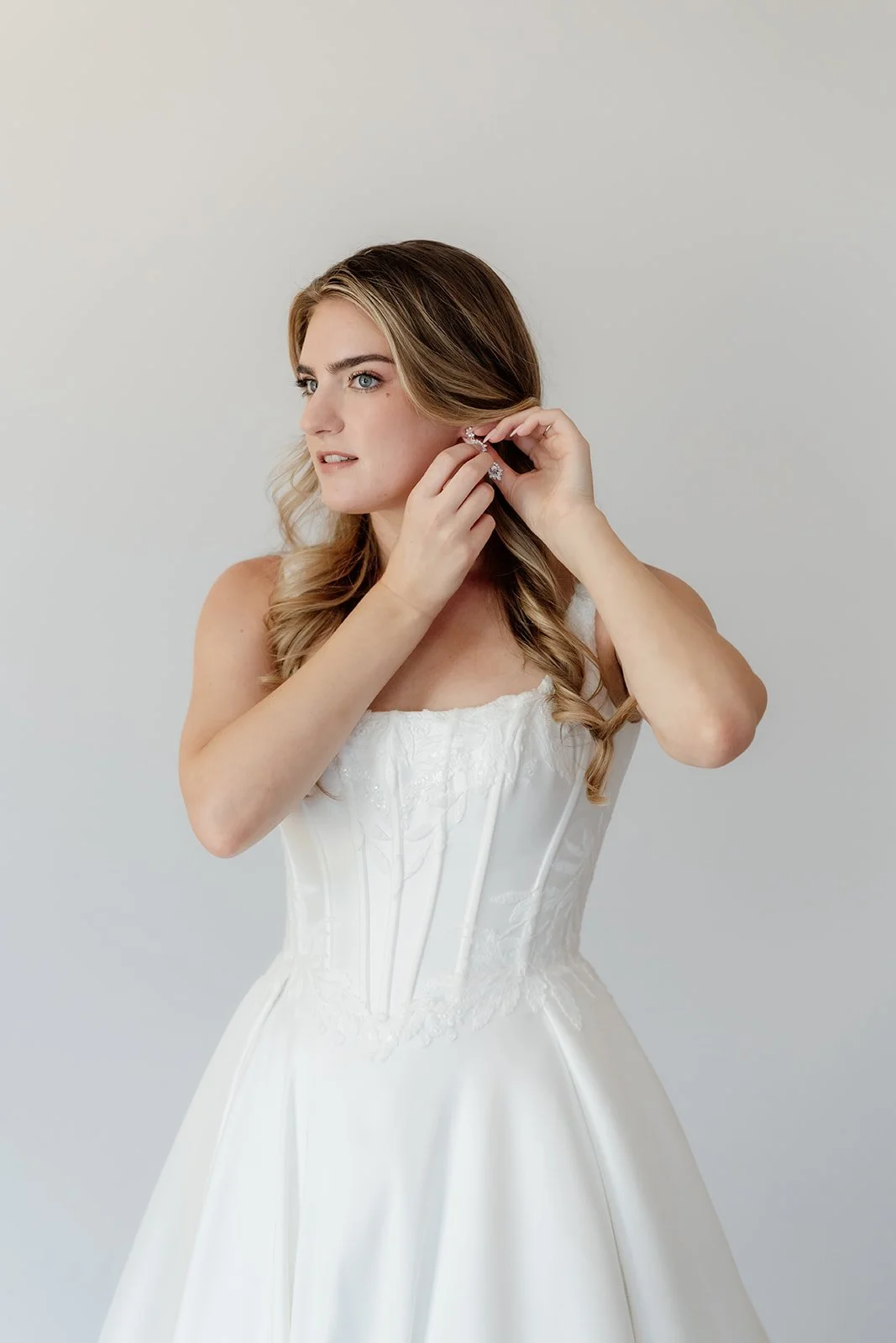 Woman in wedding dress putting on earrings against plain white background.