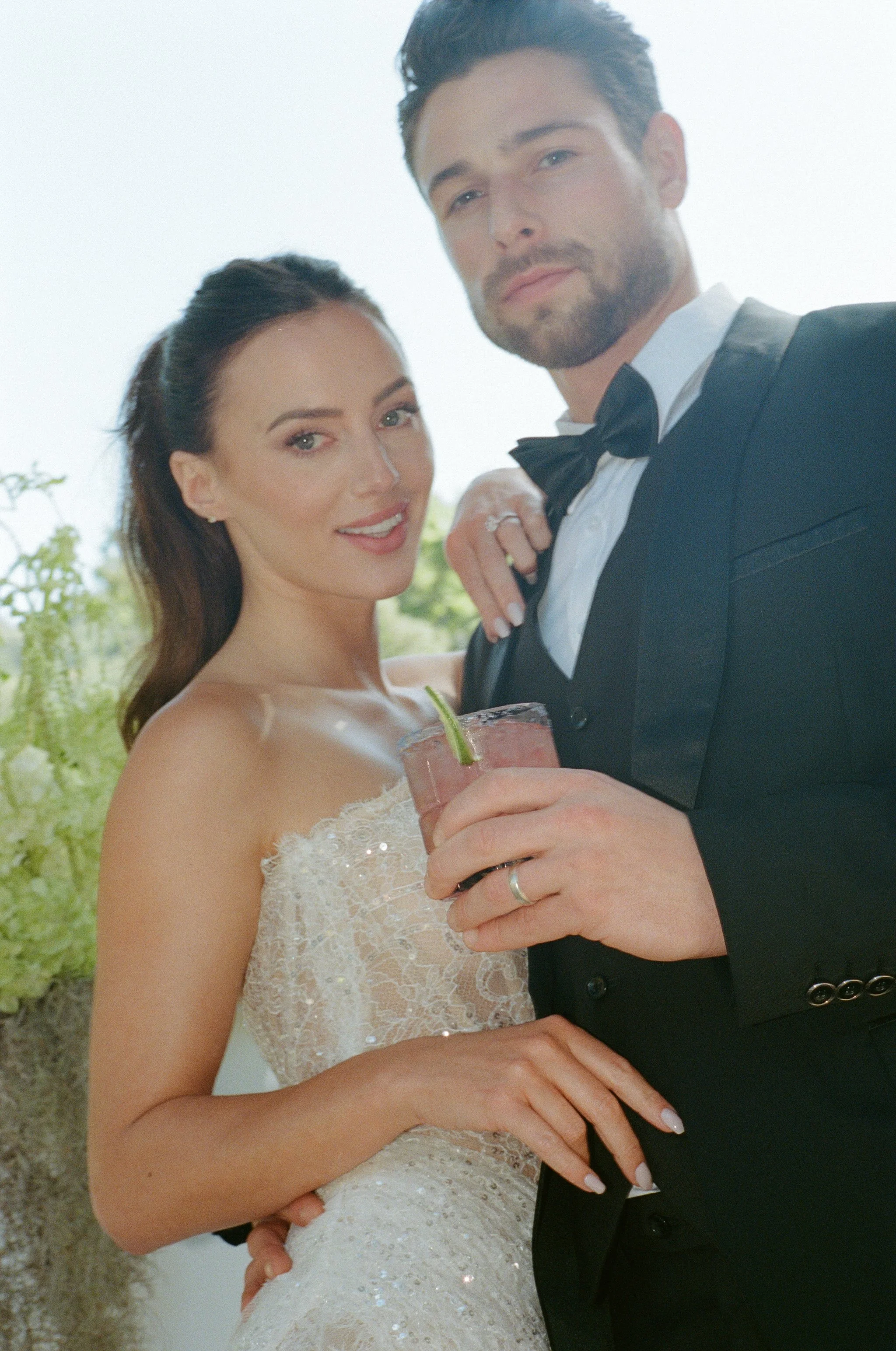 A newlywed couple dressed in wedding attire, with the bride holding a pink cocktail with a lime garnish, standing outdoors with greenery in the background.