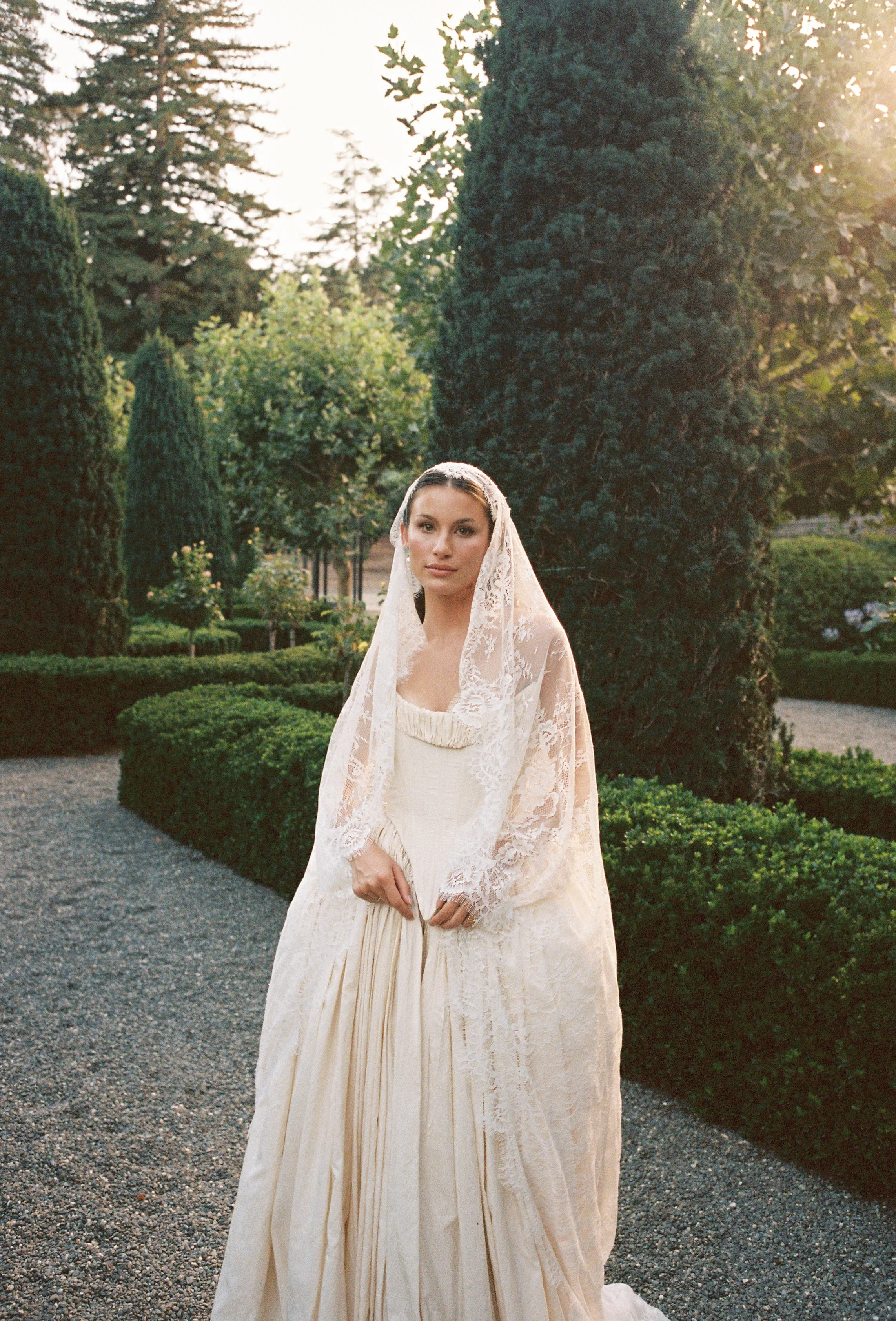 A woman in a vintage, cream-colored wedding dress with lace details, standing outdoors in a garden with tall trees and neatly trimmed bushes. The scene is illuminated by warm, natural sunlight in the evening or late afternoon.