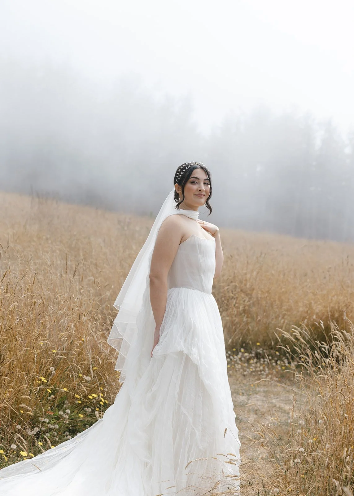A woman in a wedding dress standing in a field of tall, dry grass with fog in the background.