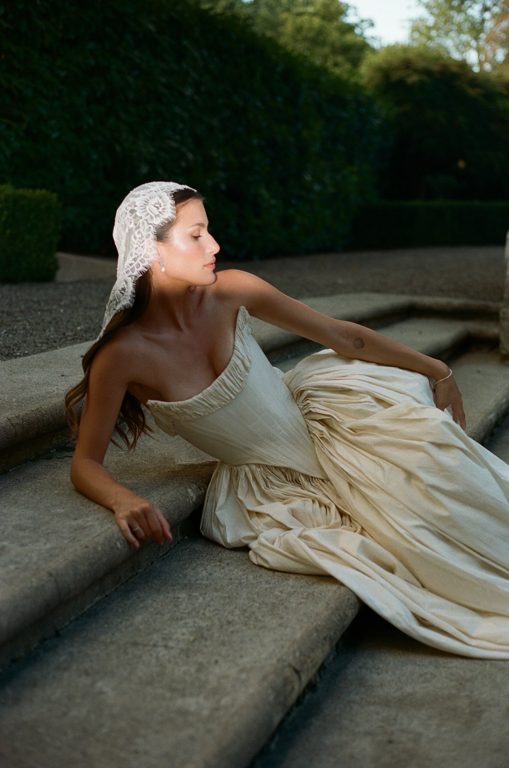 A woman in a white vintage-style dress and lace head covering sitting on a stone bench outdoors with a large hedge in the background.