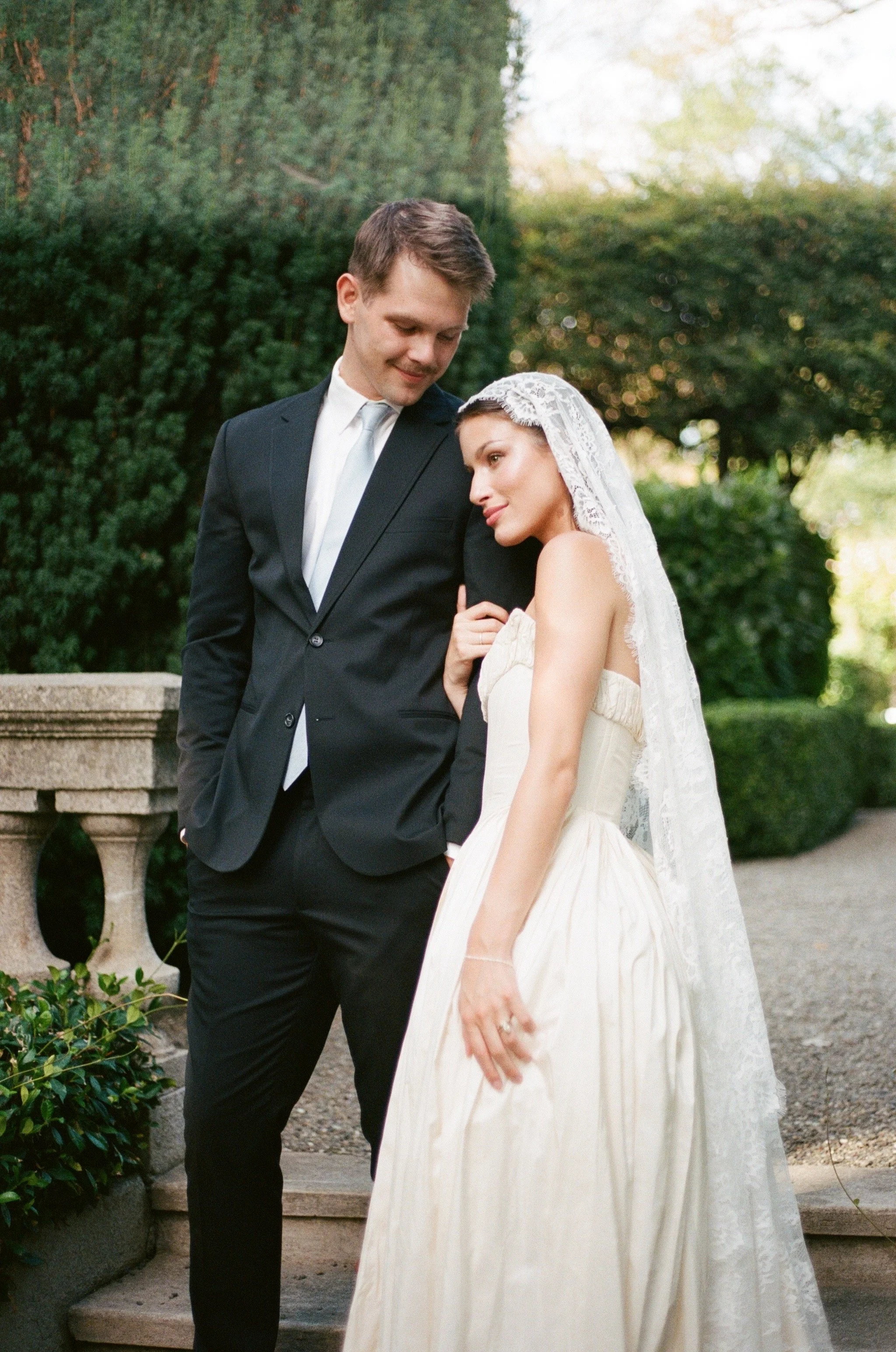 A bride and groom in wedding attire standing outdoors, with the groom in a black suit and the bride in a white wedding dress and veil, sharing a tender moment.