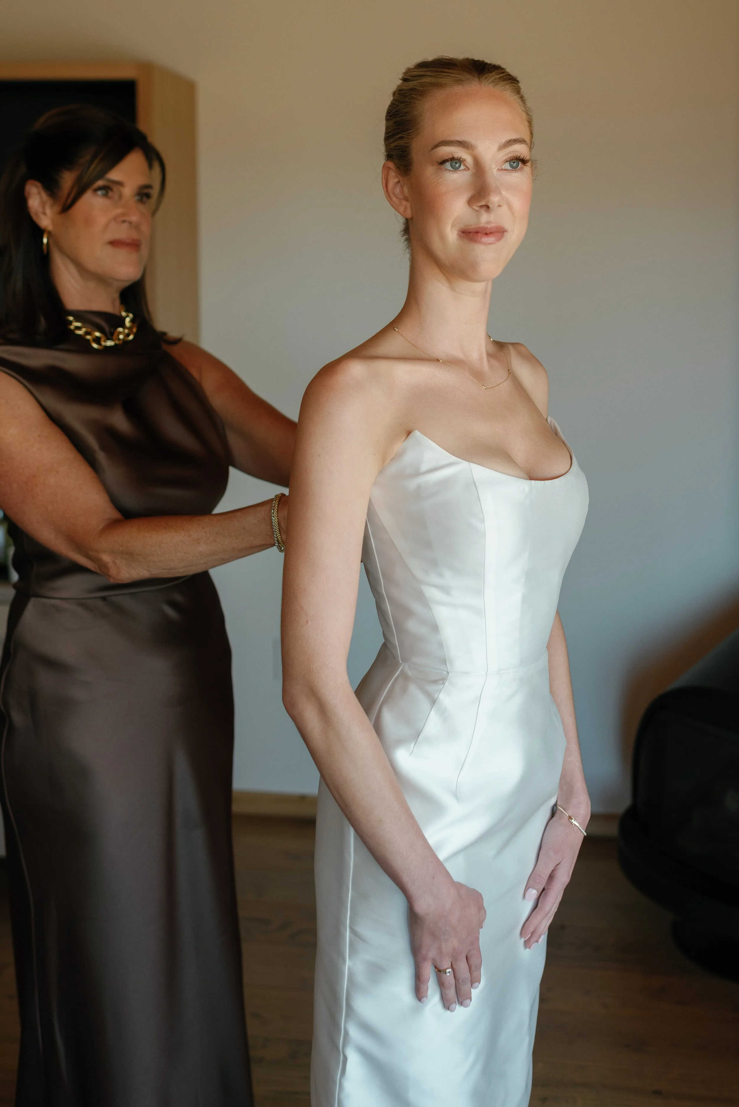 A bride with blonde hair in an updo wearing a white strapless wedding gown, standing with her hands clasped, smiling softly, as a woman in a brown satin dress adjusts her dress from behind.