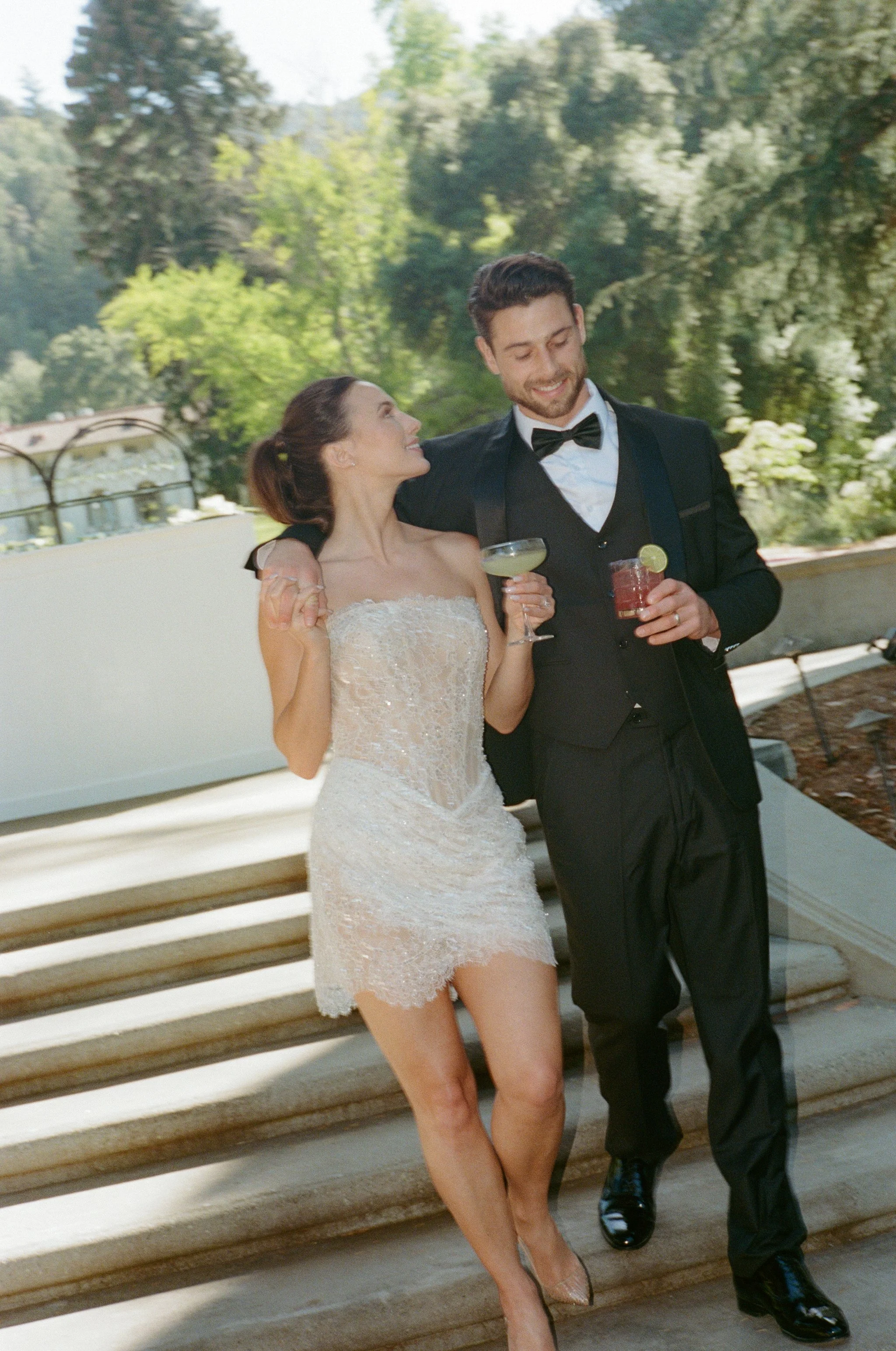 A woman in a wedding dress and a man in a tuxedo are happily standing together outdoors, holding drinks and smiling.