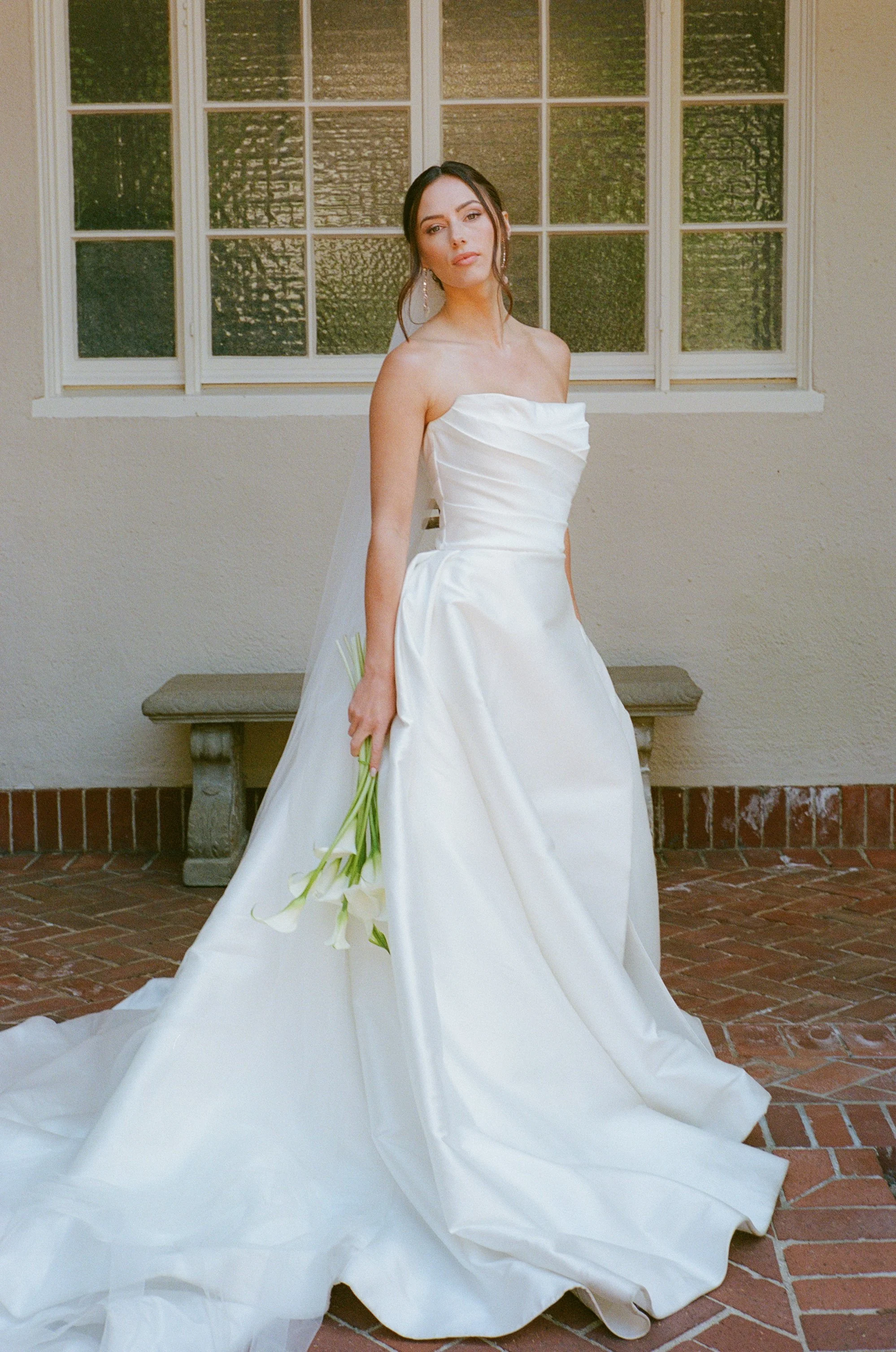 A woman in a white wedding gown standing outdoors by a window, holding a small bouquet of white flowers, with a neutral expression and dark hair styled in loose waves.