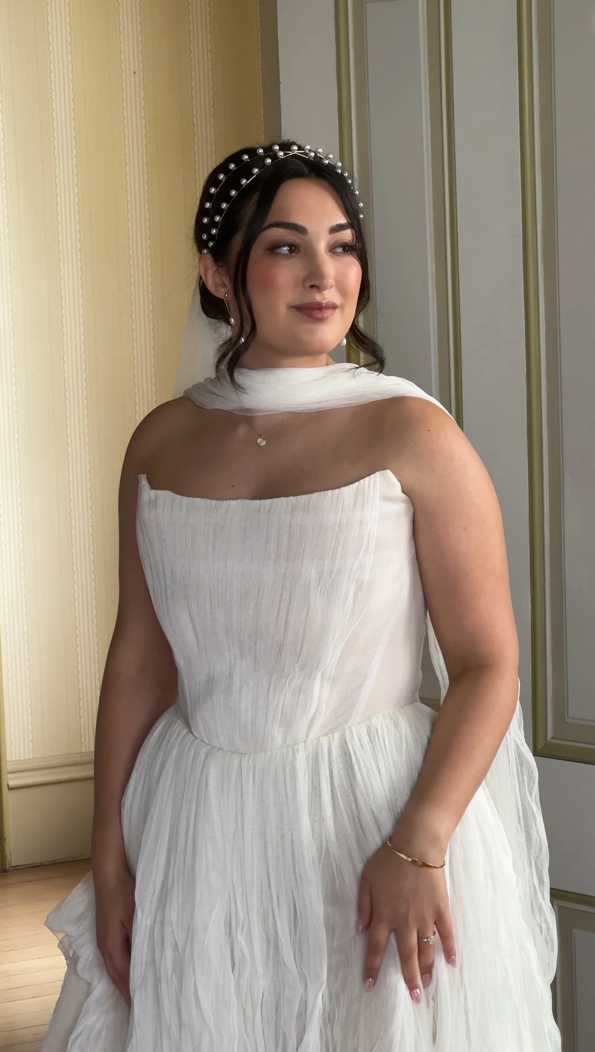 A young woman in a white wedding dress with a strapless, pleated bodice and a flowing skirt. She wears a pearl-decorated headband, a small necklace, and a bracelet. She stands indoors against a beige and gold paneled wall, with her dark hair styled i