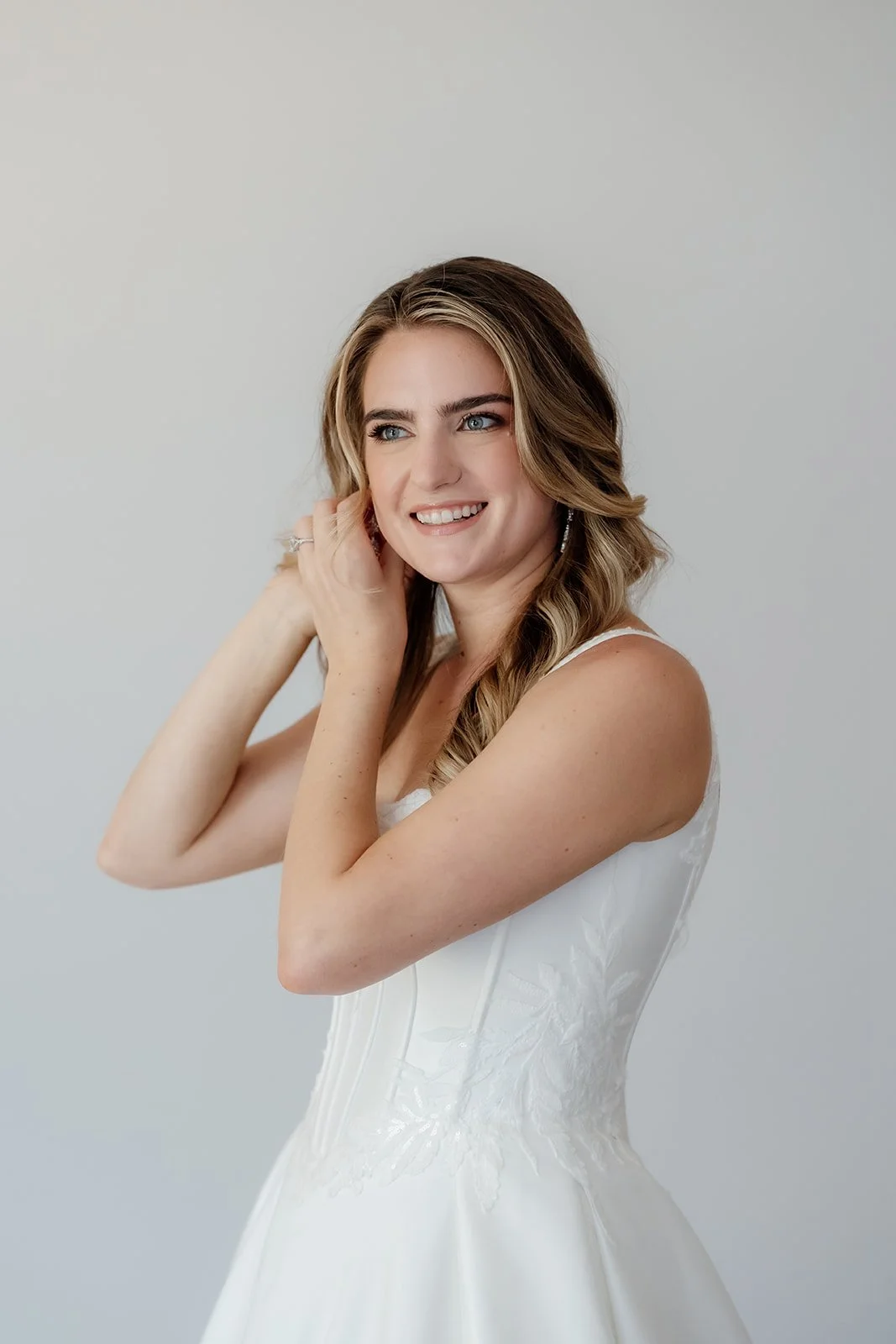 A young woman with wavy light brown hair in a white dress, smiling and touching her hair, against a plain light gray background.