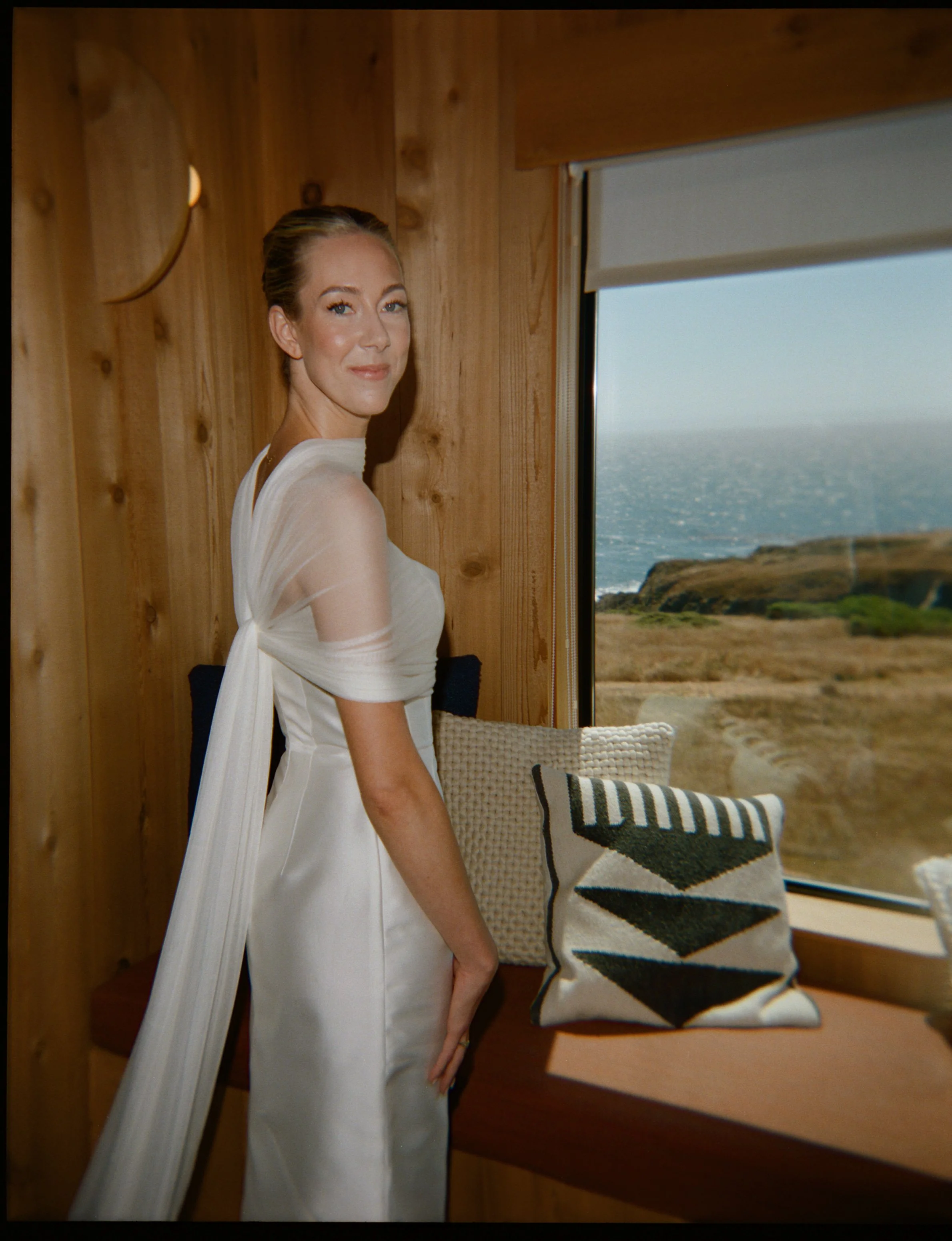 A woman in a white dress stands by a window with a view of the ocean and cliffs. She has light skin, red hair styled back, and a gentle smile.