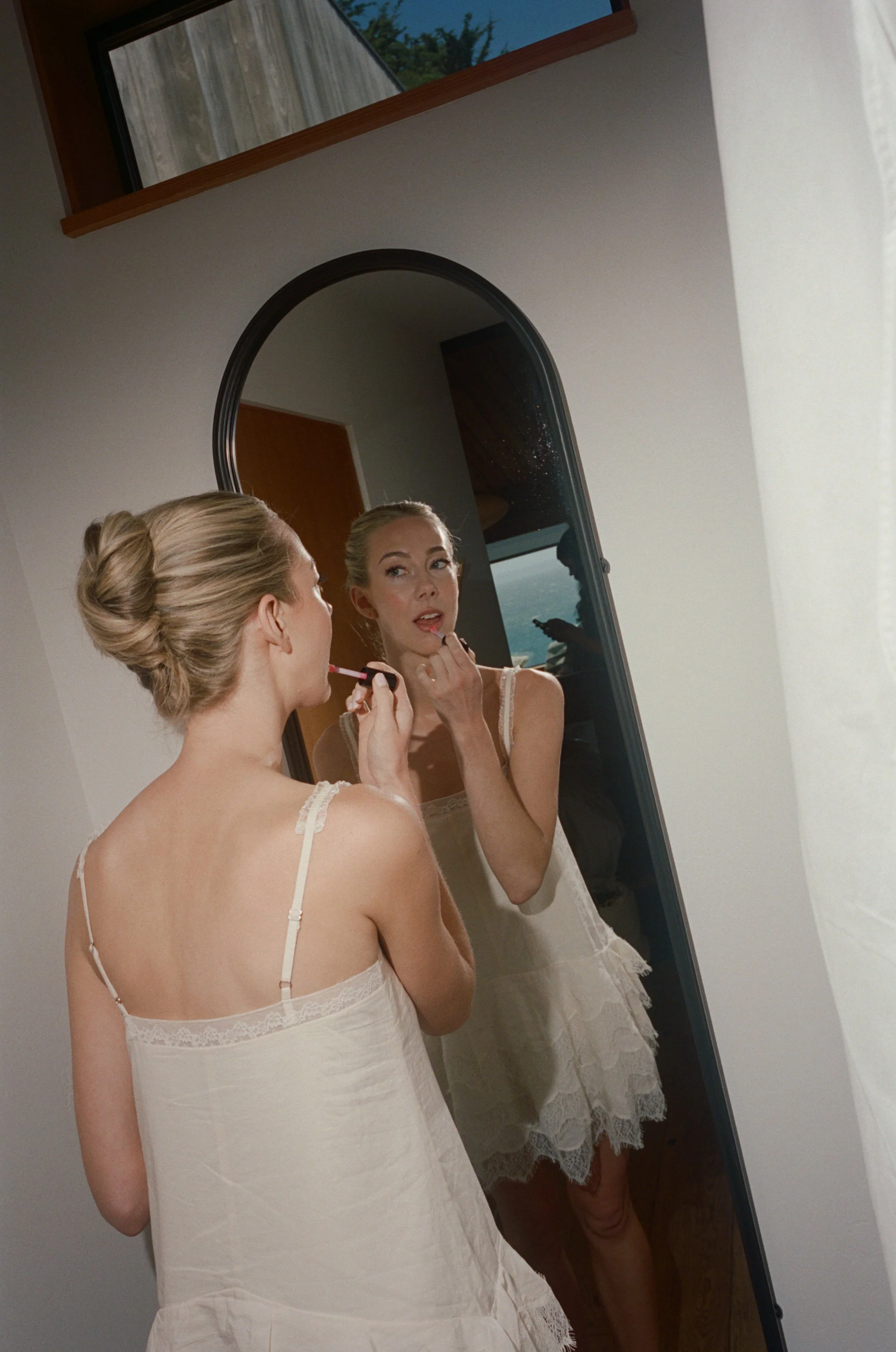 A woman with blonde hair styled in an updo, wearing a white slip dress with lace straps, is applying lipstick while looking into a mirror on a white wall. The reflection shows her face and the back of her dress. There is an arched mirror on the wall 