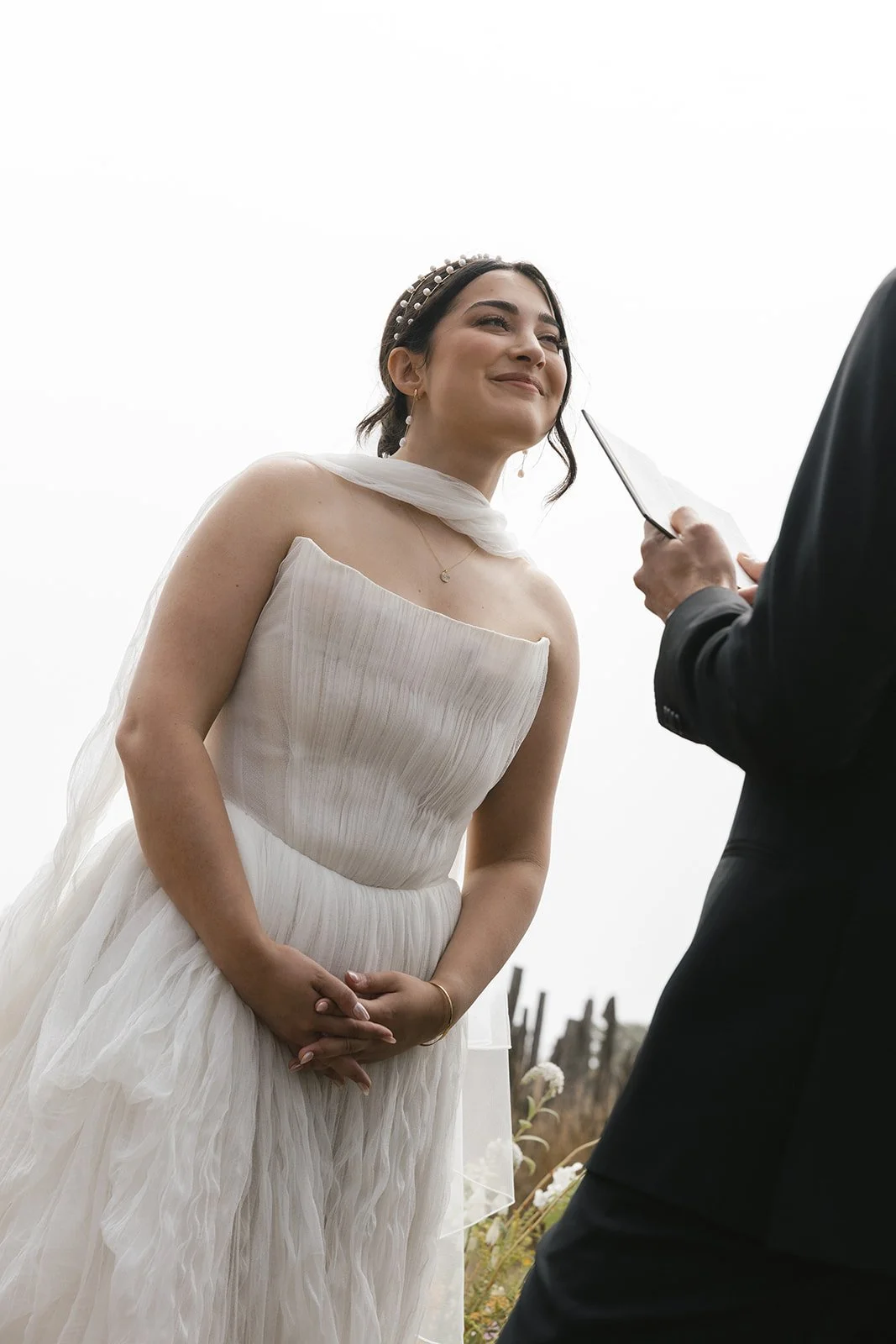 A bride in a white wedding dress standing outdoors with a slight smile, during her wedding ceremony, with a person in a black suit holding a book or binder in front of her.