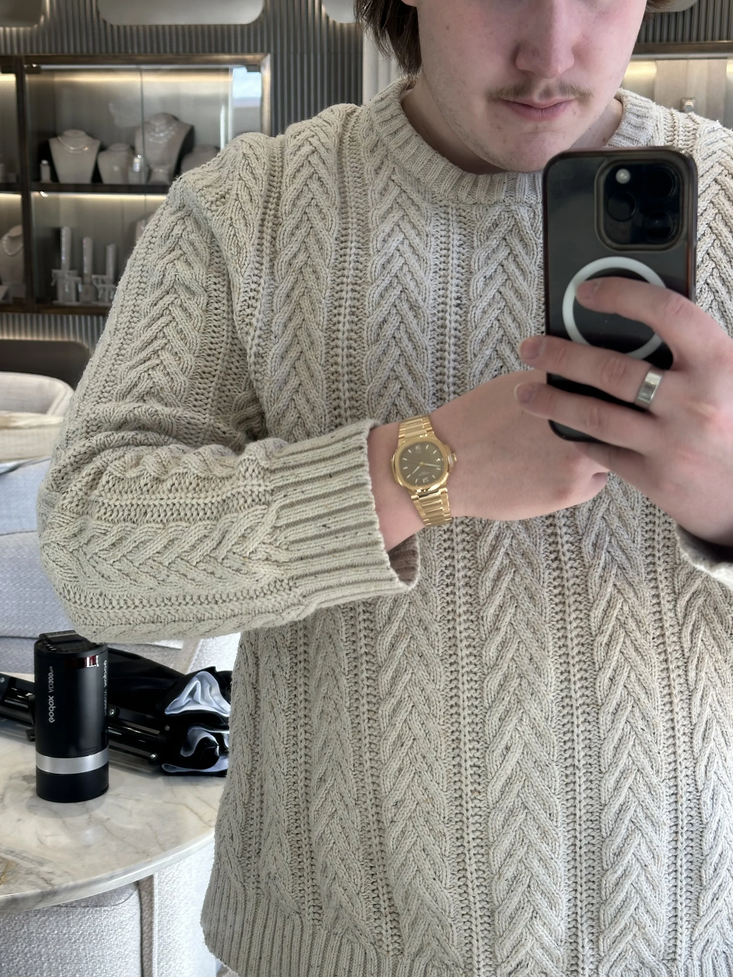 A person taking a mirror selfie wearing a beige cable-knit sweater, gold watch, and silver ring, with a display of jewelry in the background and a table with a podcast device and other items.