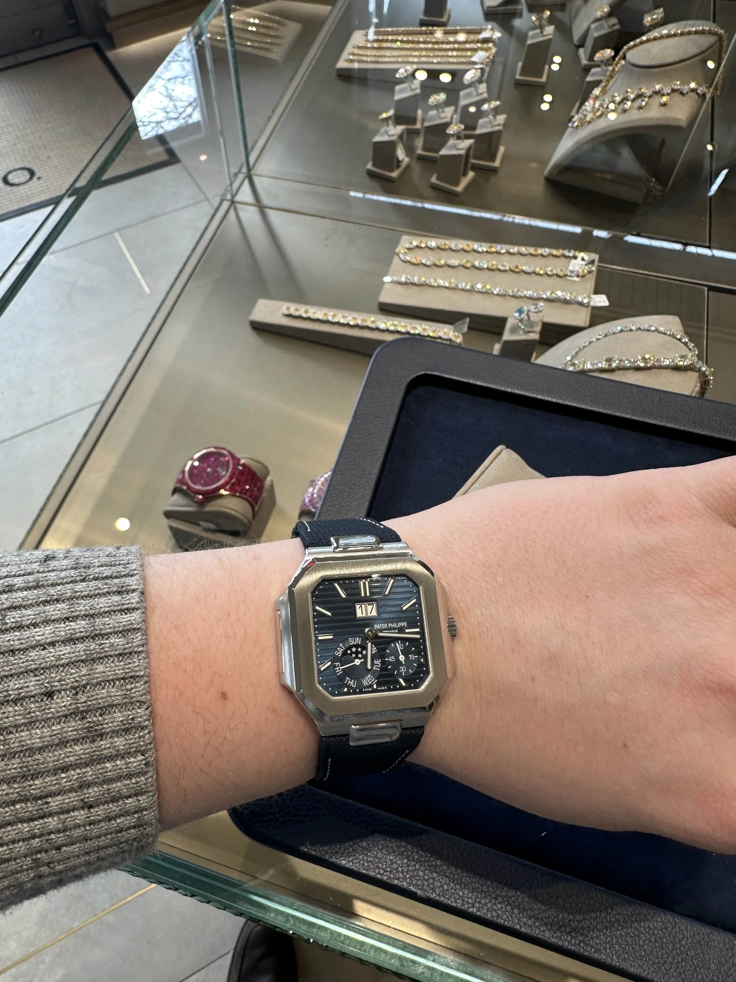 Person wearing a black and silver wristwatch with a black face showing the date, day, and small subdials, in front of a jewelry display case with diamond jewelry and accessories.