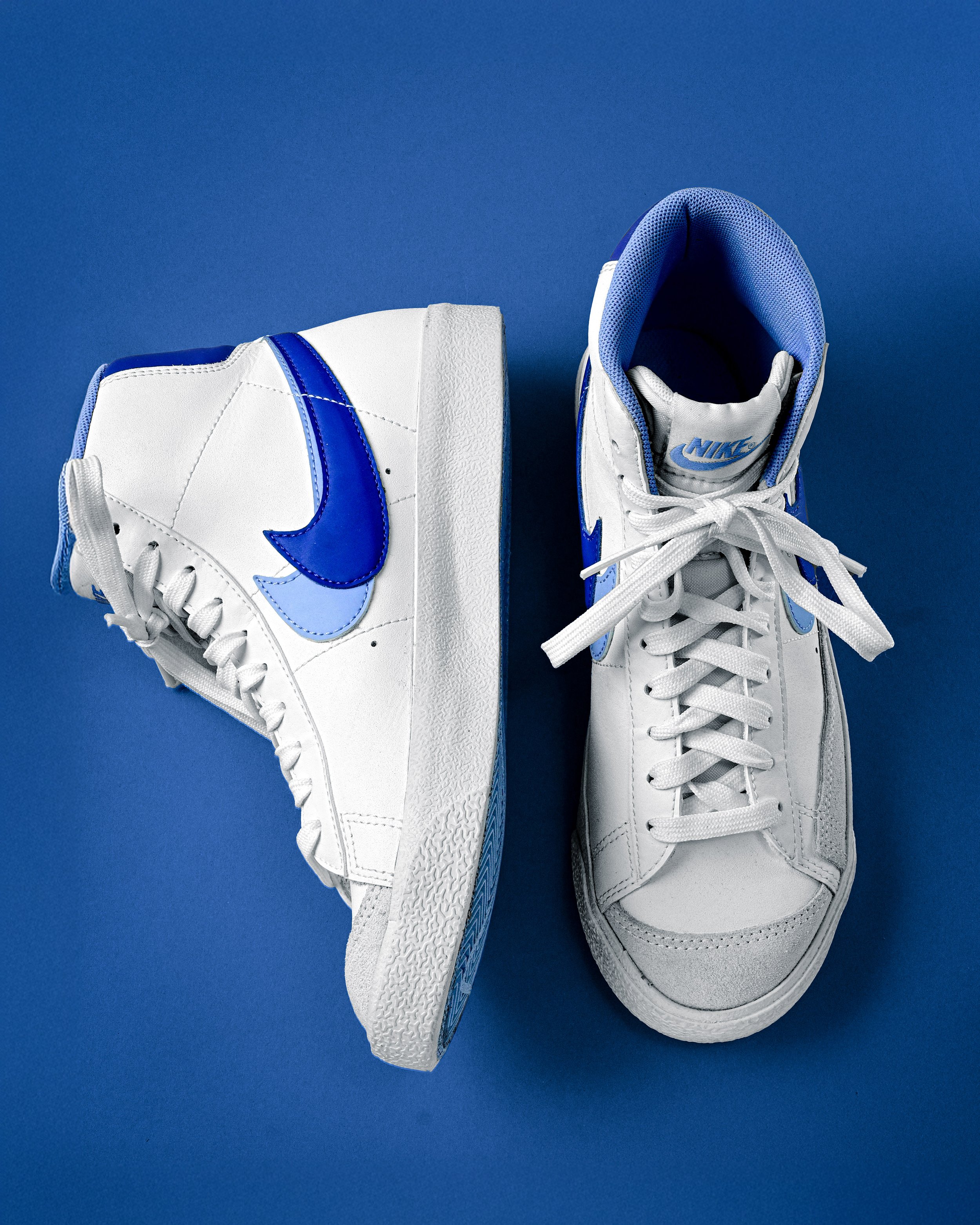 Pair of white Nike sneakers with blue accents on a blue background, one shoe standing upright and the other lying on its side.