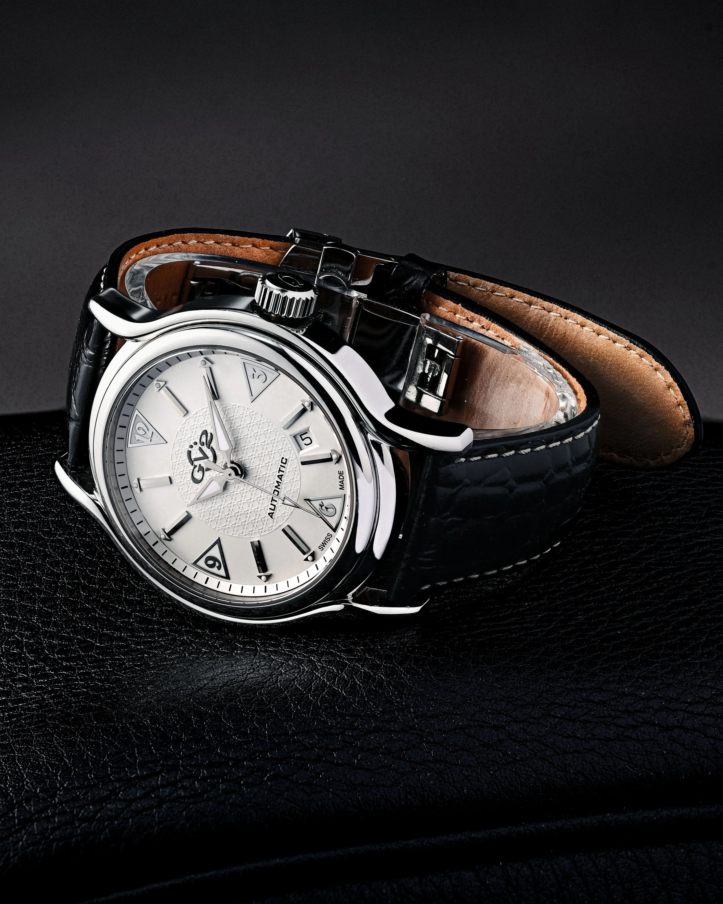 A luxury wristwatch with a silver dial, black leather strap, and classic design, resting on a black textured surface.