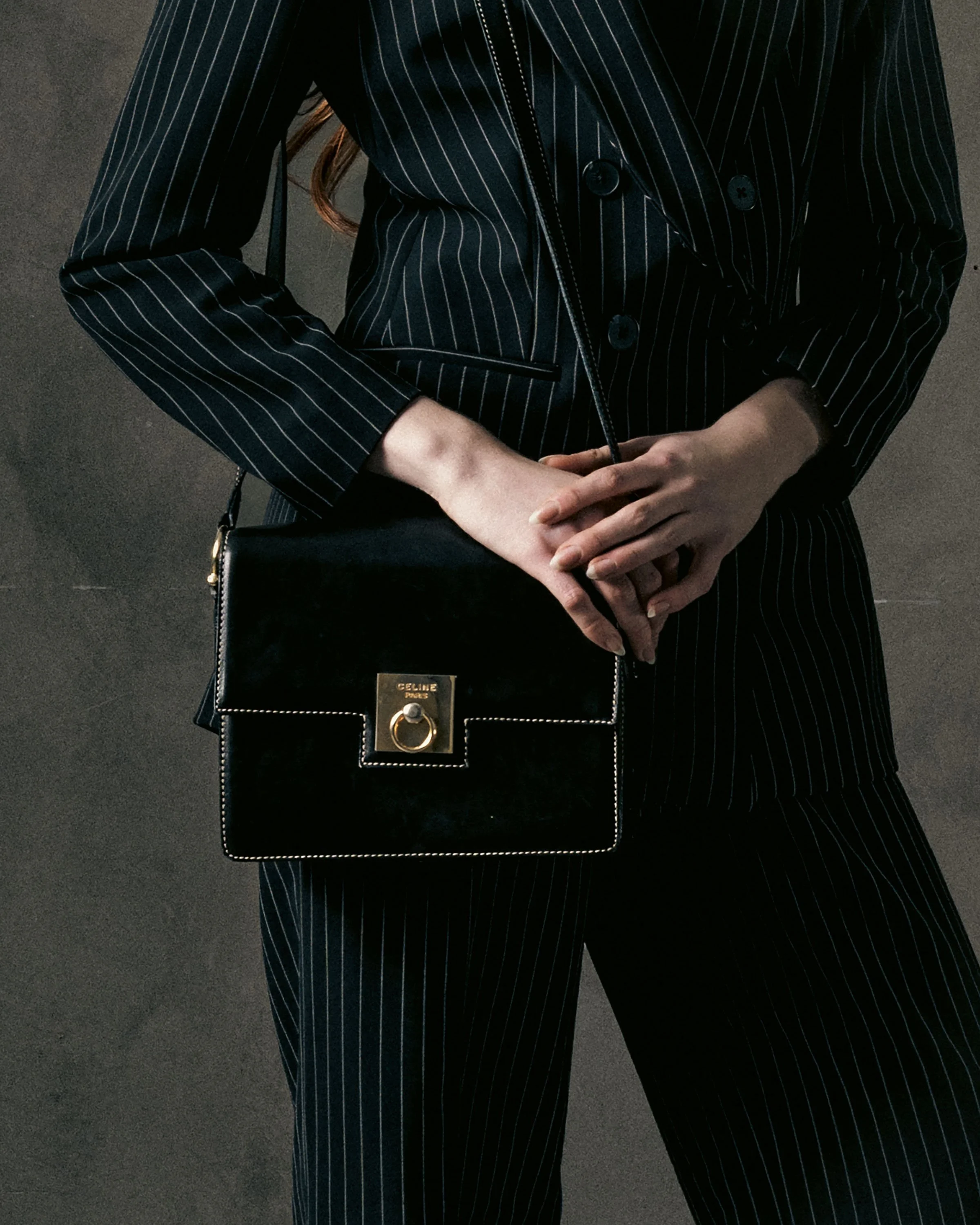 Person wearing a pinstripe blazer and matching pants, holding a black handbag with a gold clasp against a dark background.