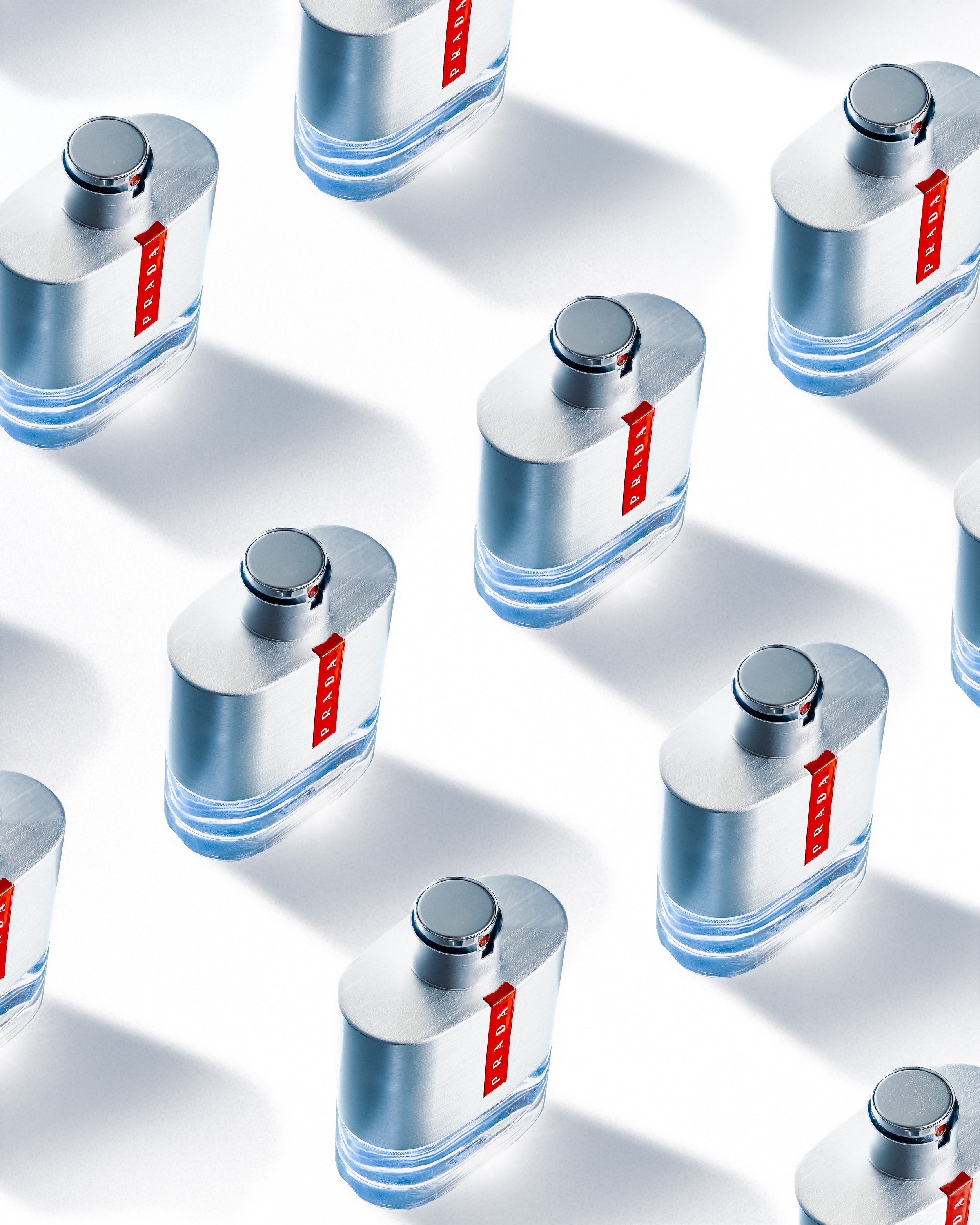 Multiple silver Prada perfume bottles with red labels arranged in a pattern on a white surface, casting shadows.