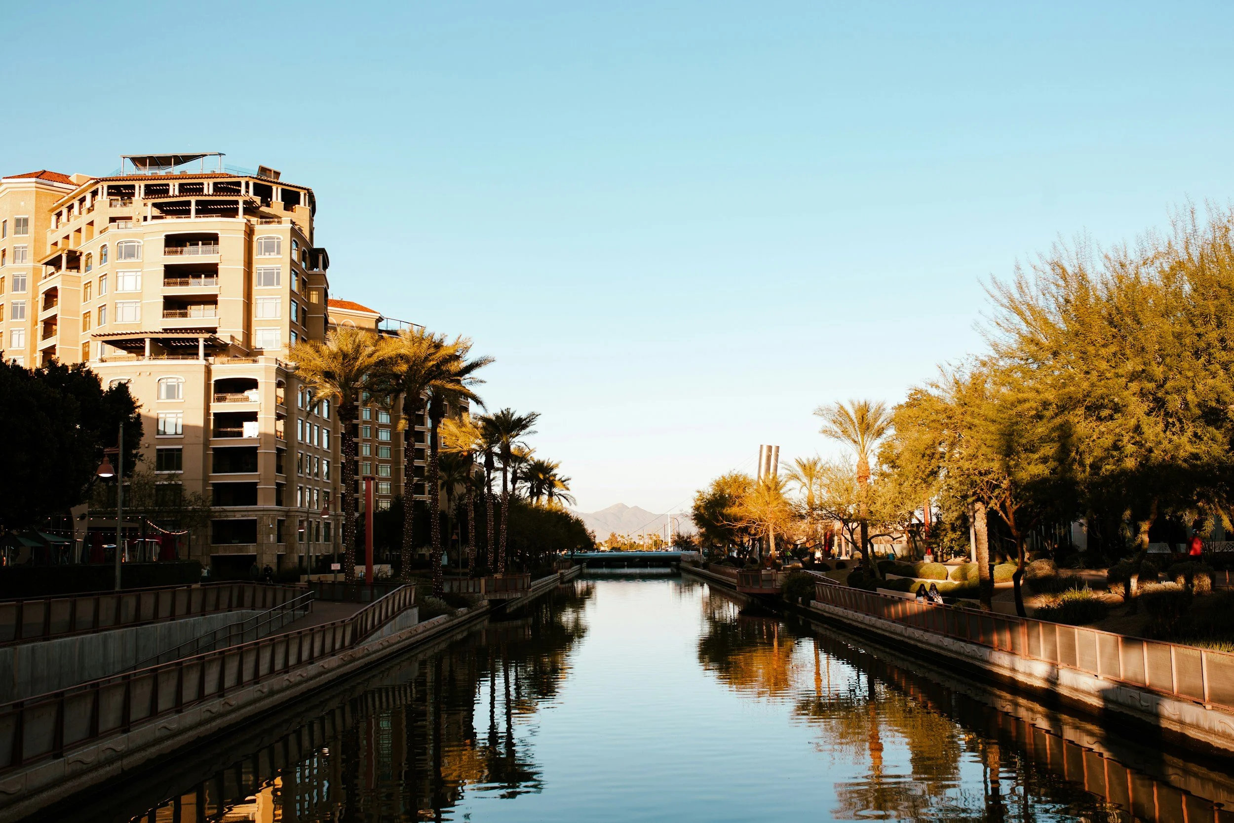 Arizona Canal at the Scottsdale Waterfront in Scottsdale, Arizona. A cityscape with a canal in the foreground, lined by palm trees and a walking path, with modern multi-story buildings on the left and trees on the right, under a clear blue sky.