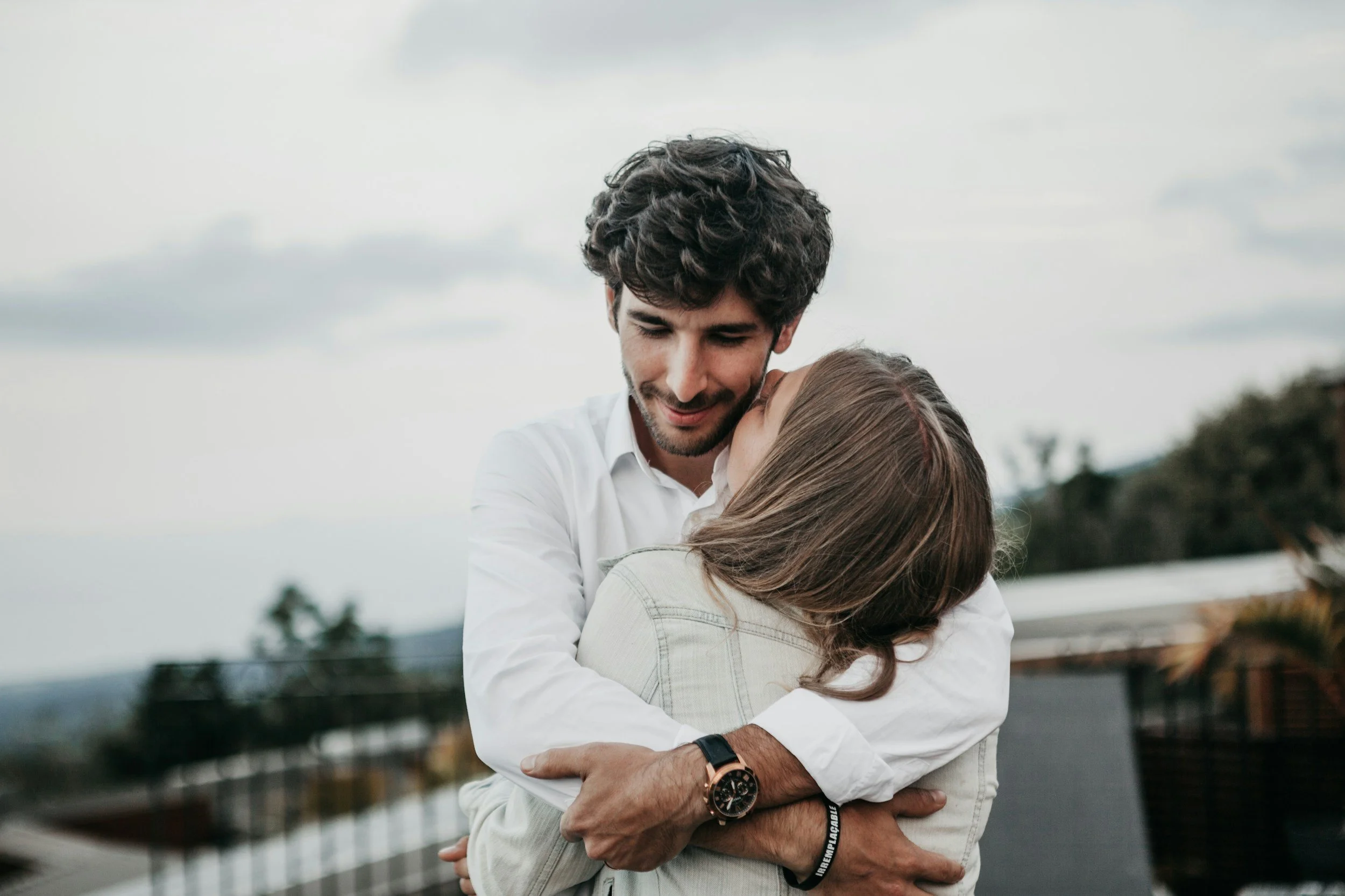 A young man and woman embrace outdoors, smiling with their eyes closed, overcast sky background, casual clothing.