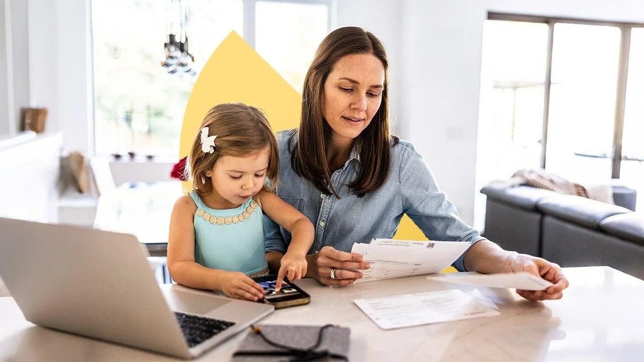 Woman and young girl at home sitting at a table with a laptop and documents, young girl using a tablet.