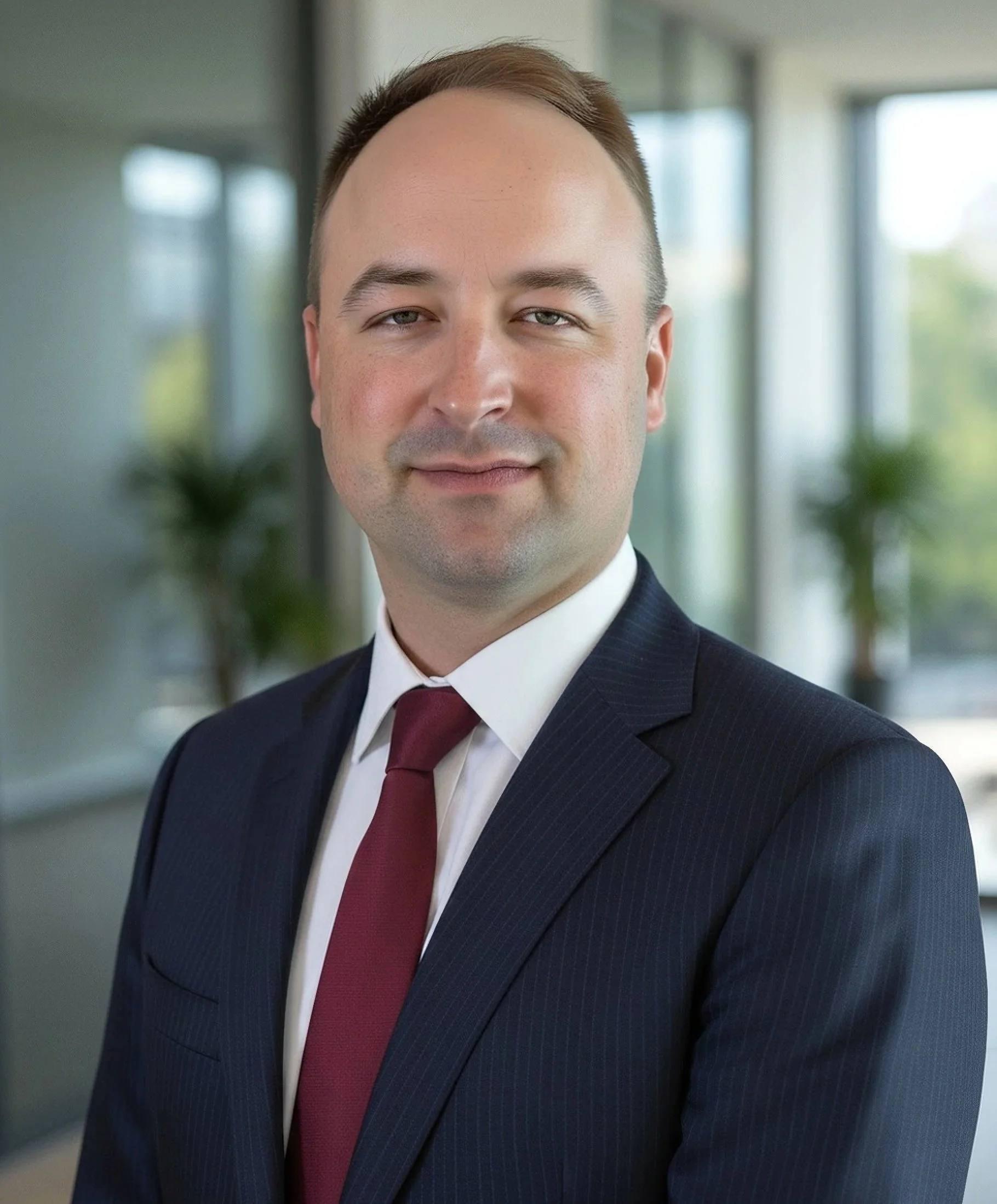 Close-up of a man with short brown hair, light skin, and blue eyes, wearing a dark pinstripe suit, white shirt, and red tie, standing indoors near large windows with blurred plants in the background.