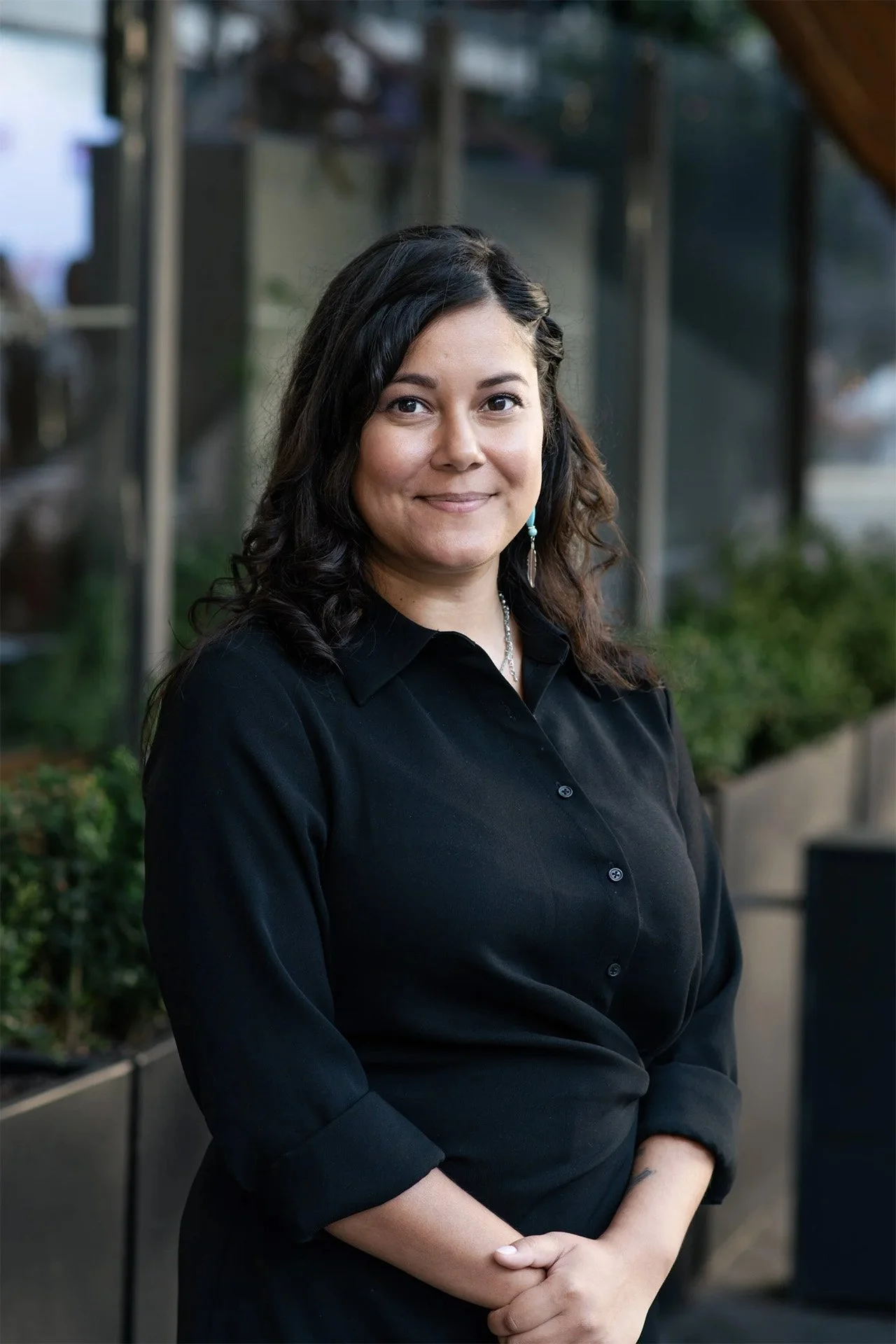 A woman with black curly hair wearing a black button-up shirt, standing outdoors in a garden or patio area with glass windows and greenery in the background.