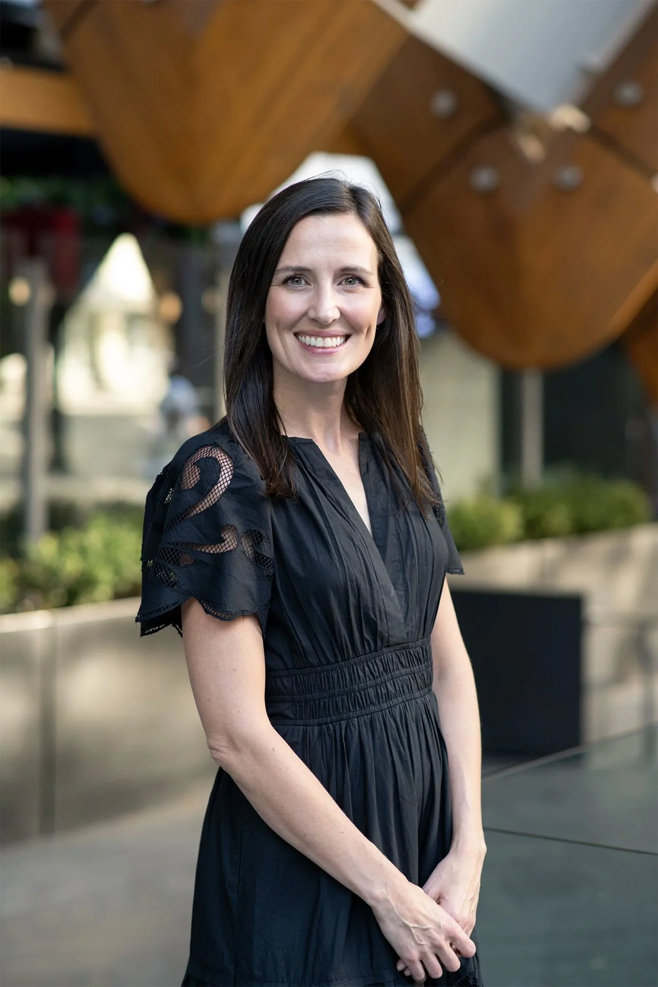 A woman with long dark hair smiling and wearing a black dress with lace details on the shoulders, standing outdoors under a large wooden sculpture.