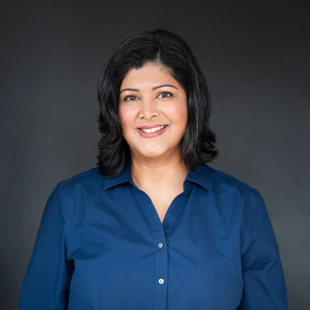 A woman with shoulder-length black hair, wearing a blue blouse, smiling against a dark gray background.
