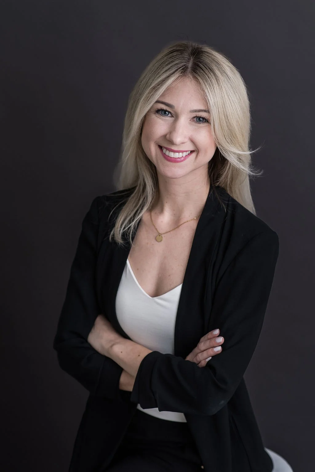 A smiling woman with blonde hair in a black blazer and white top, standing against a dark gray background.