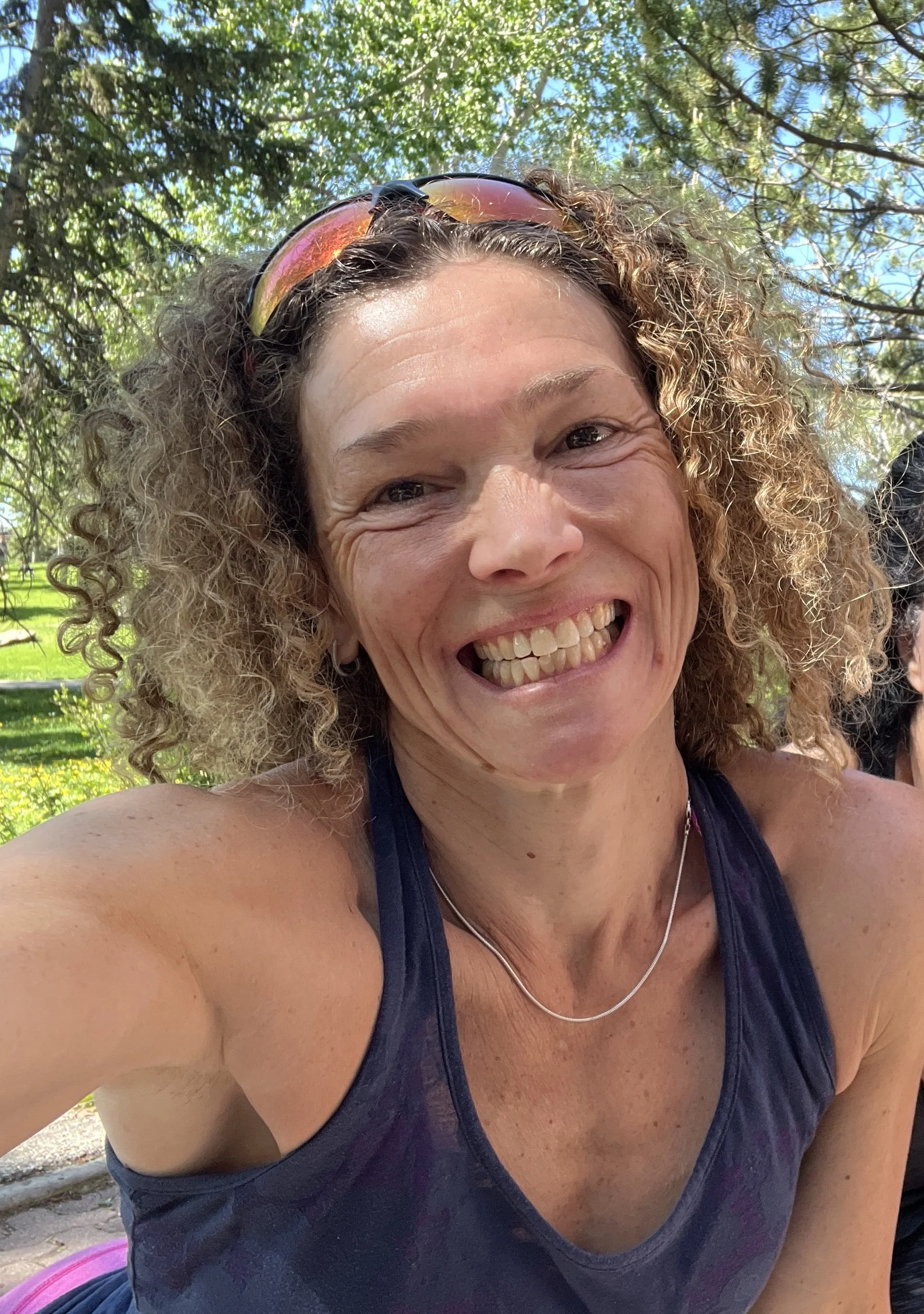 A smiling woman with curly hair wearing sunglasses on her head and a navy blue athletic top, outdoors on a sunny day with green trees and grass in the background.