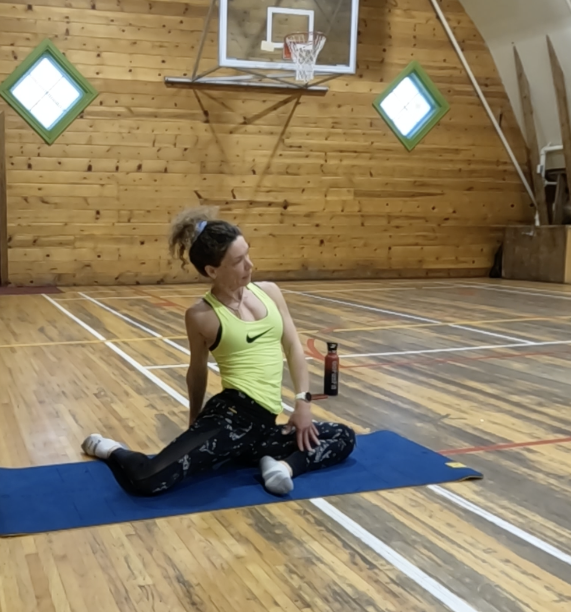 A woman with curly hair tied in a ponytail, wearing a yellow Nike tank top and black patterned leggings, is stretching on a blue mat in a wooden indoor gymnasium. A basketball hoop and a water bottle are visible behind her.
