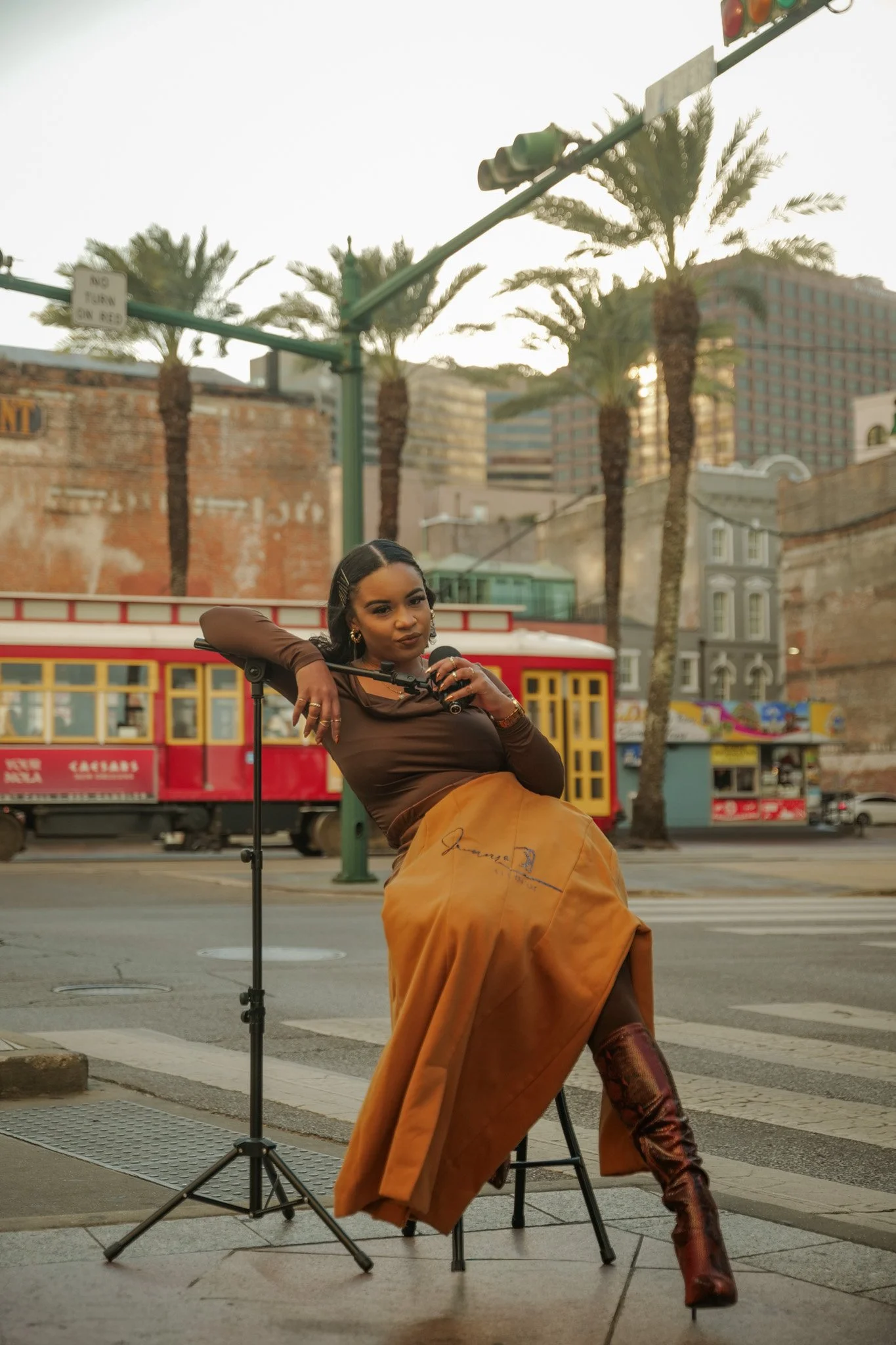A woman with dark hair and earrings sitting on a stool on a city street, holding a microphone and leaning on a music stand with palm trees and a vintage trolley bus in the background.