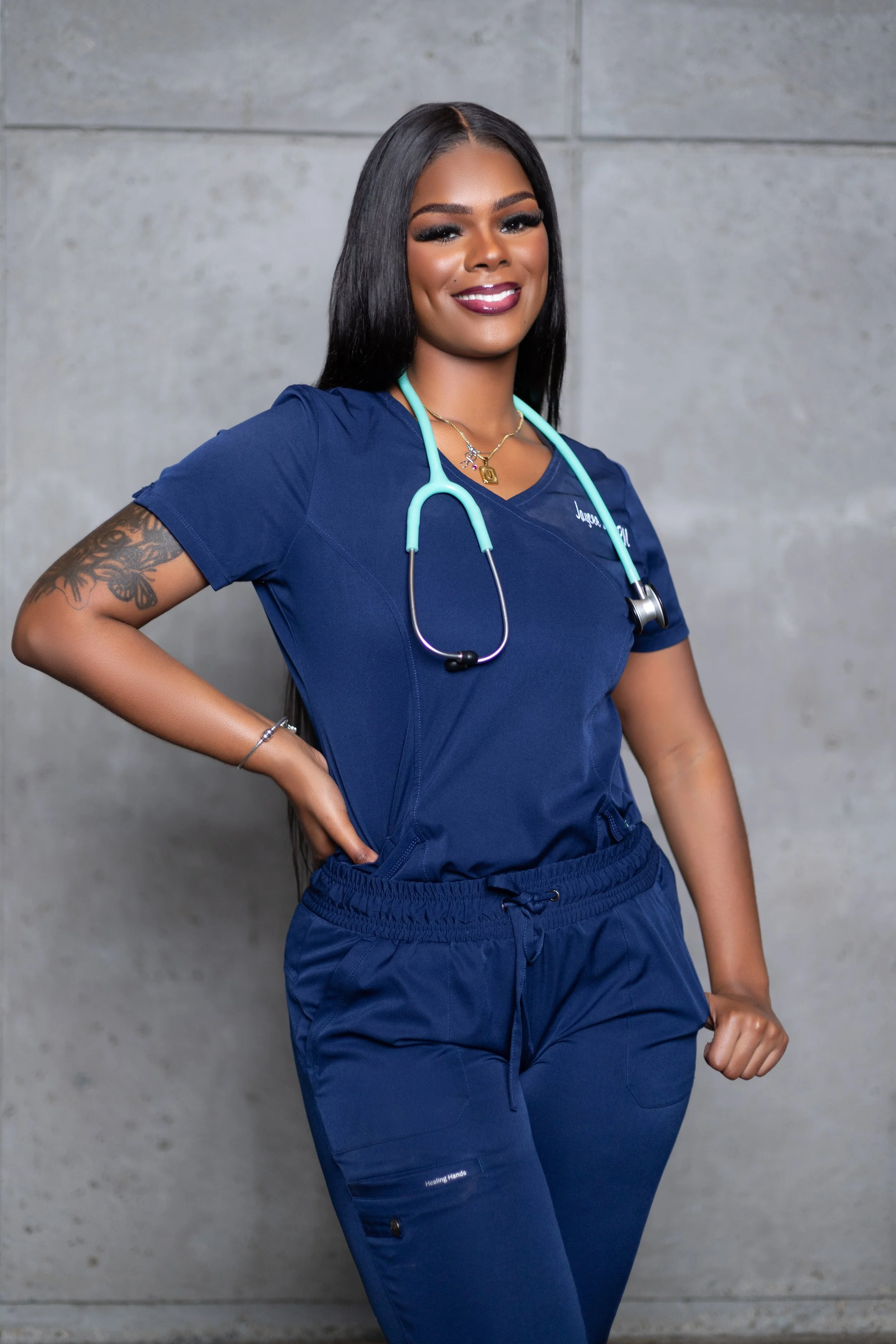 A confident nurse in navy scrubs stands against a concrete wall, smiling with her hand on her hip, wearing a stethoscope around her neck.