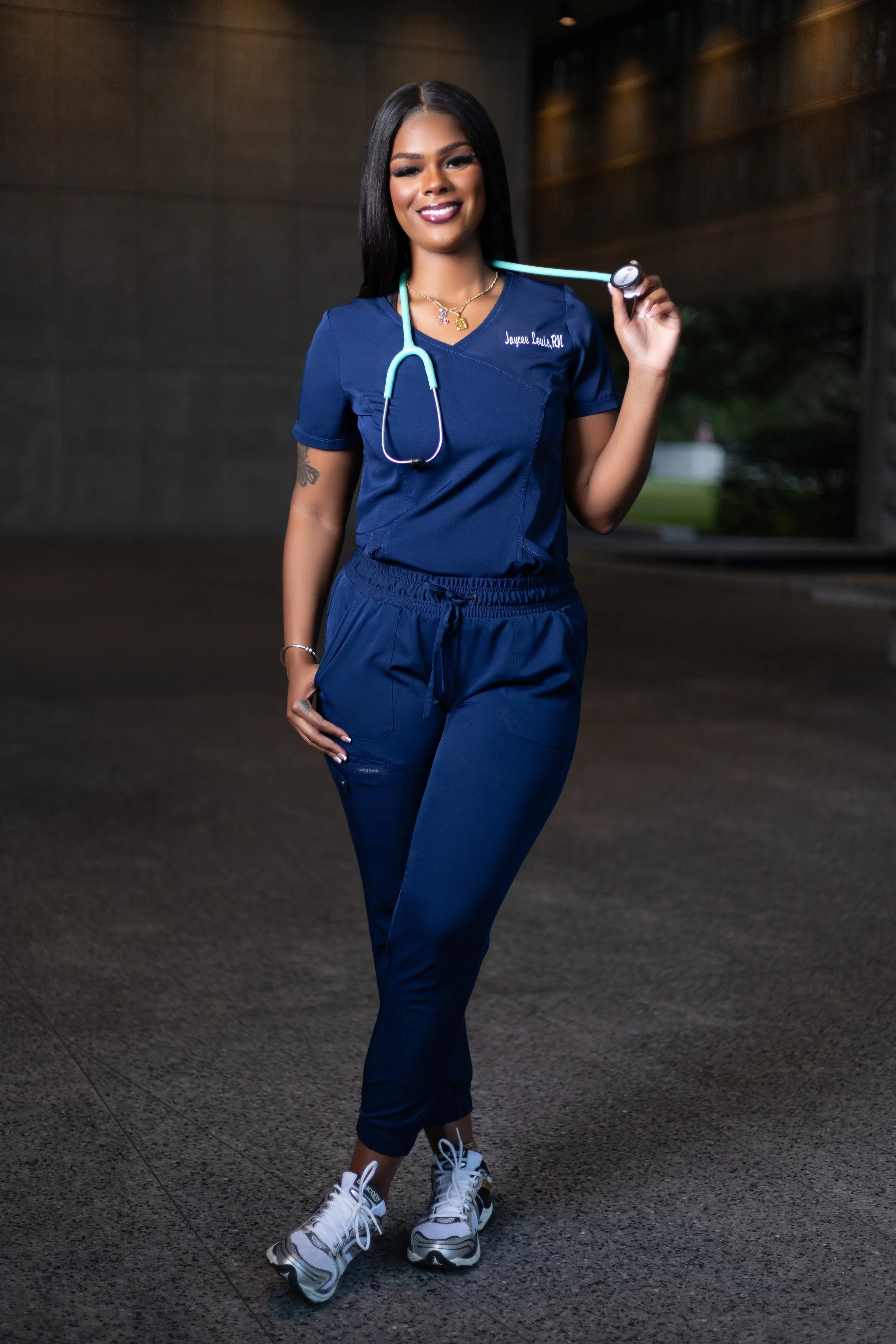 Young female nurse wearing navy scrubs, holding a stethoscope, standing outside at night with a confident smile.