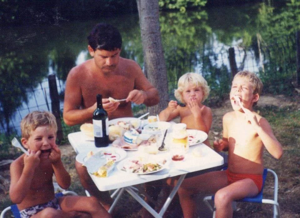 Old photo of Alice, her 2 older brothers and Dad, sat having a dinner at a French campsite, next to a river.